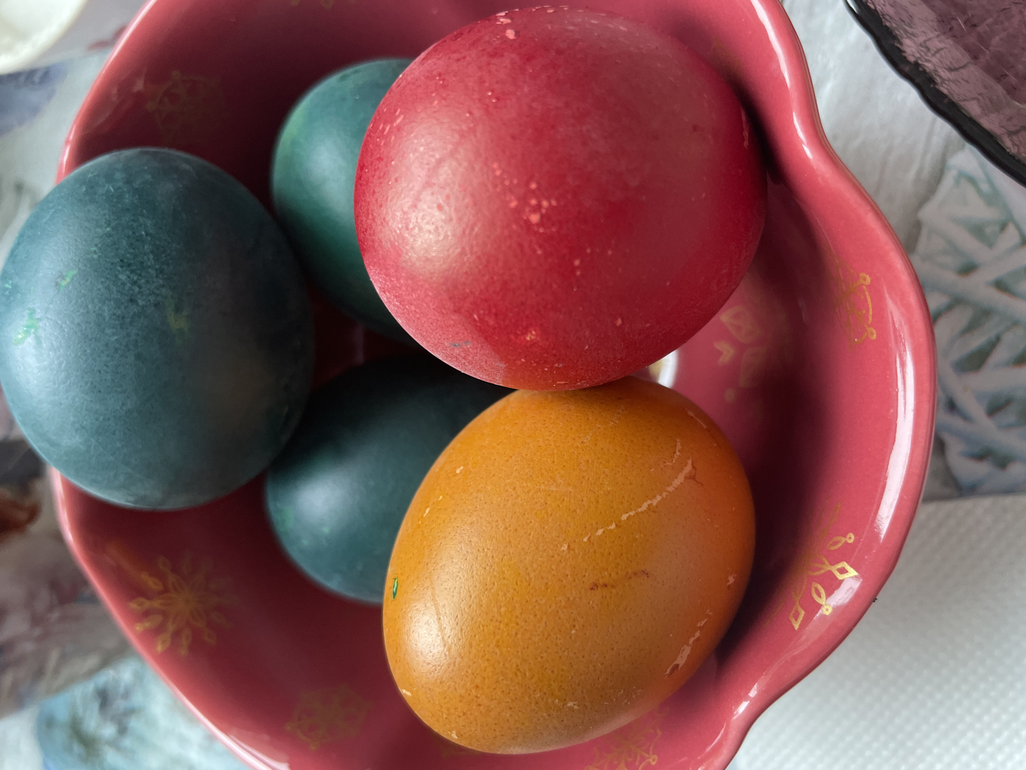 A top-down close-up shows a decorative pink bowl containing five dyed Easter eggs. The eggs are colored red, orange, and two shades of blue. The bowl's interior is decorated with golden patterns. The location is the city of Chaikovsky, Russia.