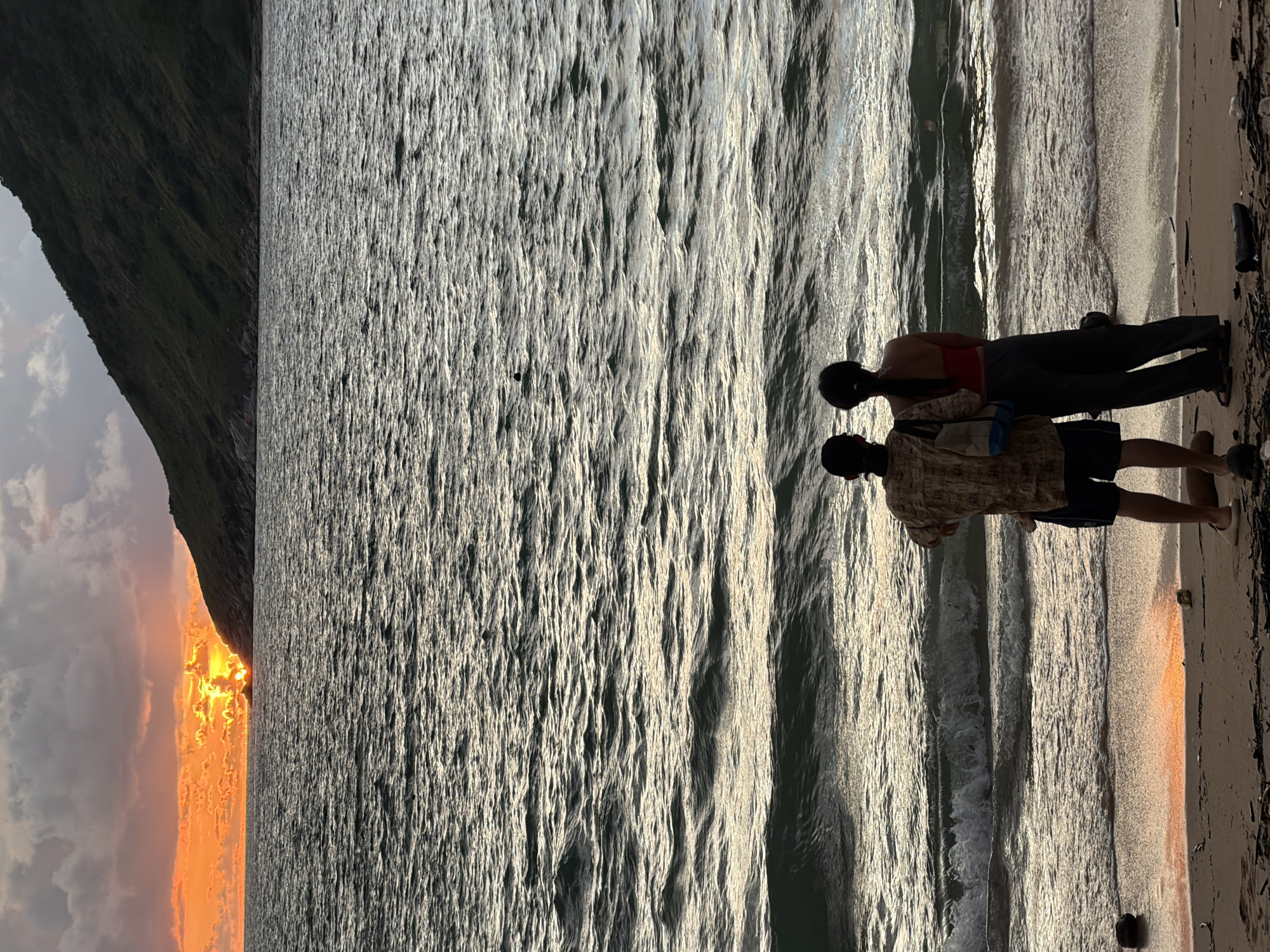The media file shows two people standing on a beach, facing the ocean. One person, wearing a red top and dark pants, stands slightly behind and to the right of the other person. The second person, wearing a patterned shirt and dark shorts, is facing away from the camera, looking towards the water. Both individuals appear to be in silhouette against the bright water and sand.
The scene is a beach at sunset, with the sun casting a warm, golden glow on the water and sand. The ocean is calm, with gentle waves lapping at the shore. The sky is partly cloudy, with some dramatic orange and pink hues from the setting sun visible on the left side of the image, juxtaposed against a dark, steep cliff face. The overall mood is peaceful and serene, suggesting a moment of quiet contemplation or enjoyment of nature's beauty.
There is no visible text in the media file. The location cues, such as the presence of a steep cliff face meeting the sea and the warm sunset light, are consistent with a tropical coastal setting. Given the context provided, this scene is likely in Rawai, Thailand, a popular beach destination known for its scenic sunsets.