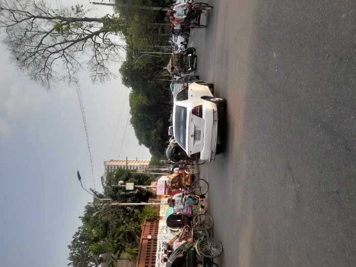 The image shows a street intersection in Dhaka, Bangladesh, at latitude 23.732 and longitude 90.395. There are several people walking and riding bicycles. A white sedan is stopped in the intersection. There is a large tree in the background. The street is paved with asphalt. There is a building on the left side of the street. The sky is overcast. There are several power lines and streetlights visible. A rickshaw with two passengers is on the left side of the road. The street is relatively busy.