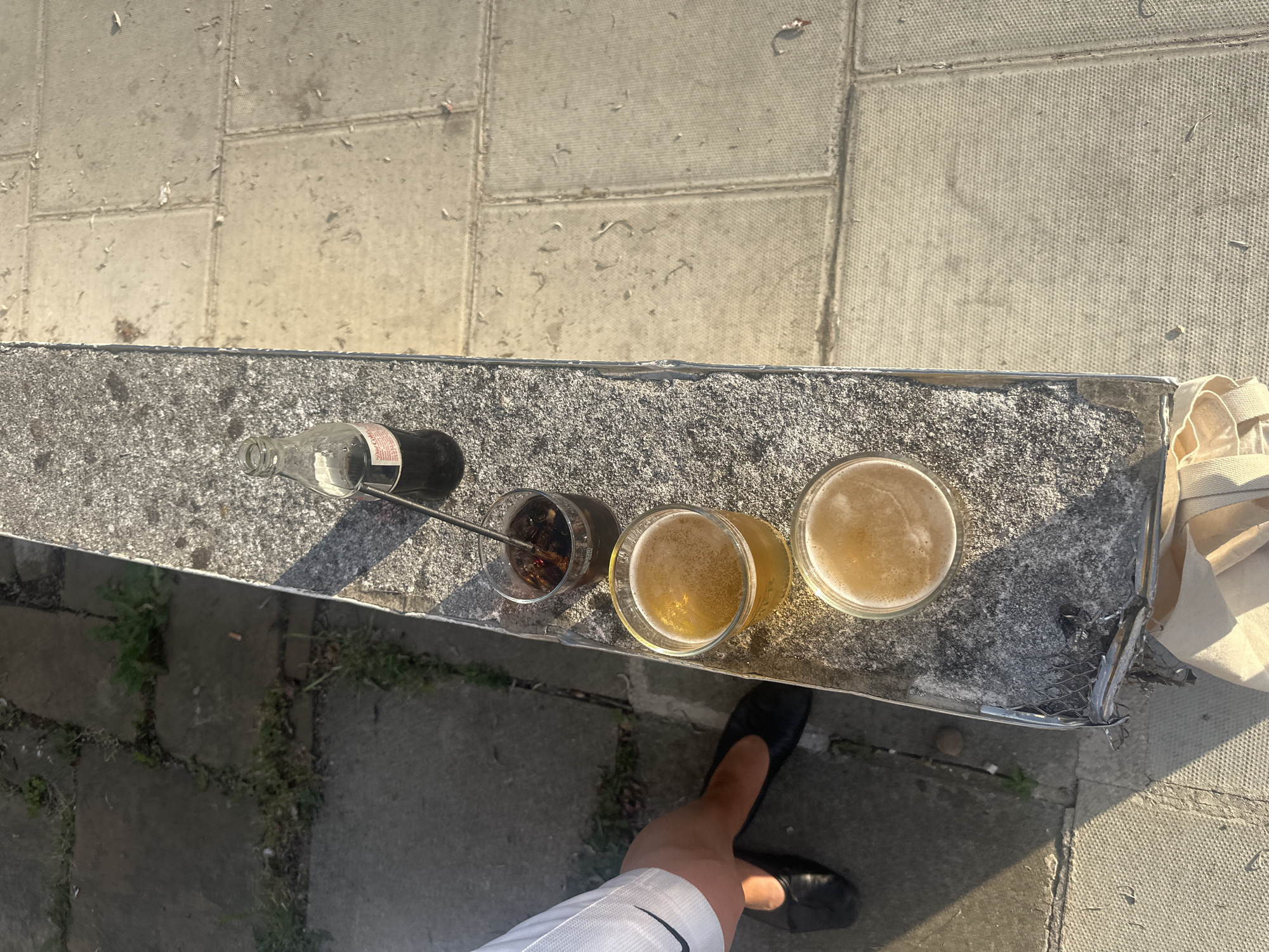 The photograph, taken from an elevated perspective, depicts a portion of a weathered, possibly public, concrete structure or bench, set against a backdrop of paving stones. On the concrete surface are several drinking vessels: an empty glass Coca-Cola bottle lying on its side, a glass containing a dark-colored liquid with what appear to be ice cubes and a straw, and two glasses filled with beer. A person's leg, clad in white shorts with a visible Nike logo, and a black shoe are visible in the foreground, suggesting that the person is standing or walking adjacent to the concrete surface. To the right, a white fabric bag rests on the concrete surface. The setting appears to be outdoors, with sunlight casting shadows across the scene. The location may be in London, United Kingdom, but there are no specific features to confirm this.