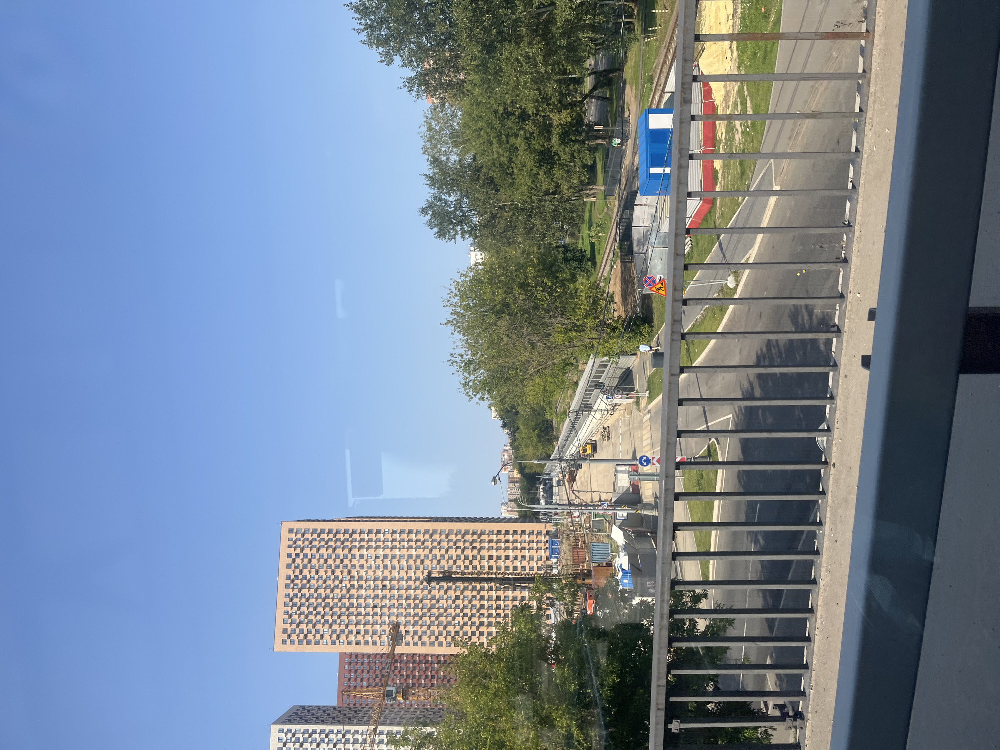 The image captures a view of a street under construction in Moscow, Russia. In the background, several tall residential buildings are visible, some under construction with cranes. The foreground features a metal railing, suggesting the photo was taken from a bridge or elevated walkway. Below, a road curves through the scene, with construction materials, barriers, and some parked vehicles. A blue and white construction trailer sits beside a red and white structure on the side of the road. Trees and a grassy area line the road on the right, with railway tracks running parallel to it. The sky is clear and blue, indicating a sunny day.
