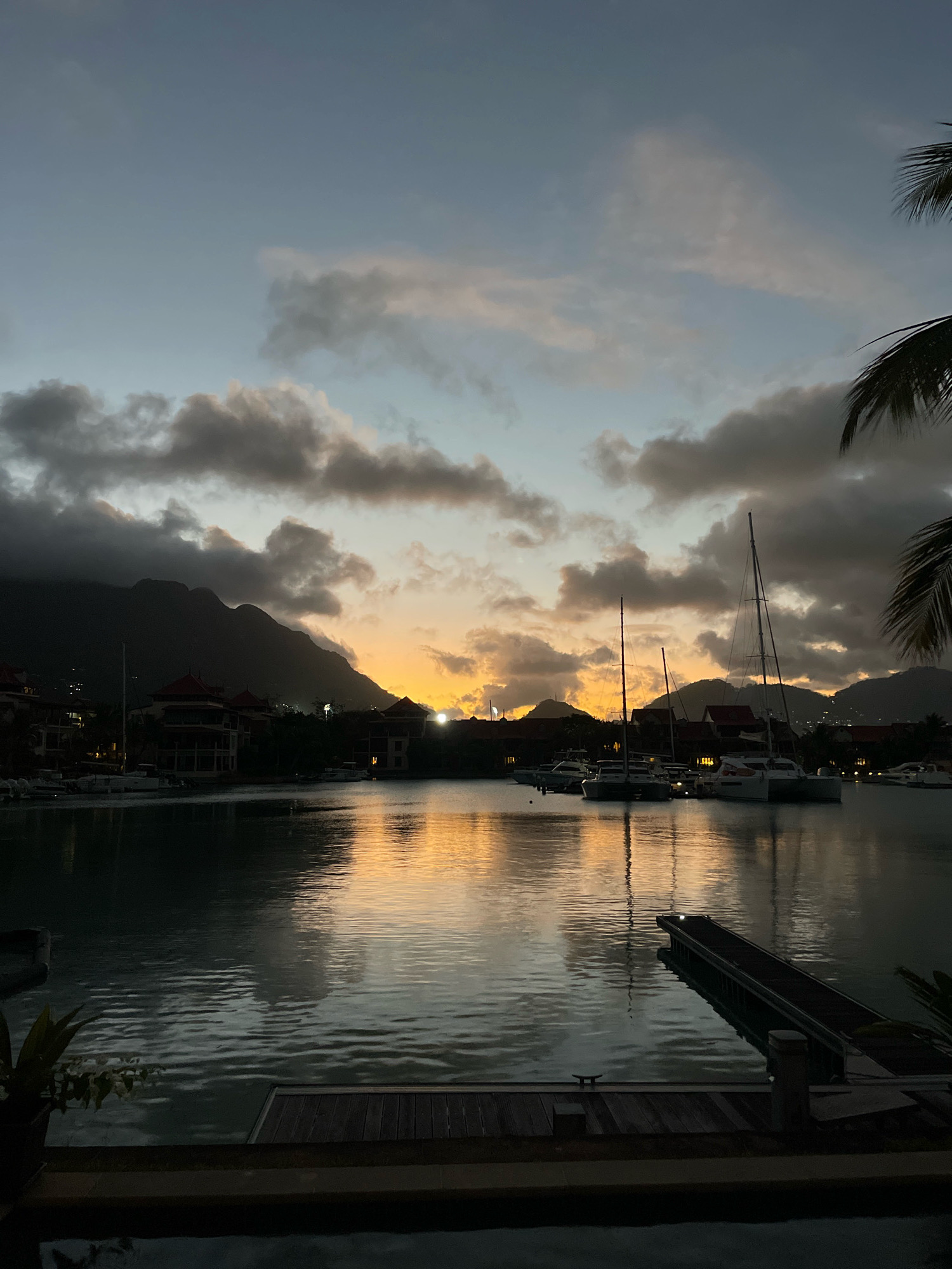 A twilight view of a harbor in the Eden Island area of Victoria, Seychelles, features calm water reflecting the sky and the silhouettes of mountains in the background. Several sailboats and catamarans are docked in the harbor. Buildings with red-tiled roofs line the shore, and palm fronds are visible in the foreground and right side of the frame. The sky is a gradient of dusky blue and orange, with clouds that are illuminated by the setting sun. A wooden pier extends into the water on the right, and another wooden deck is in the lower foreground.