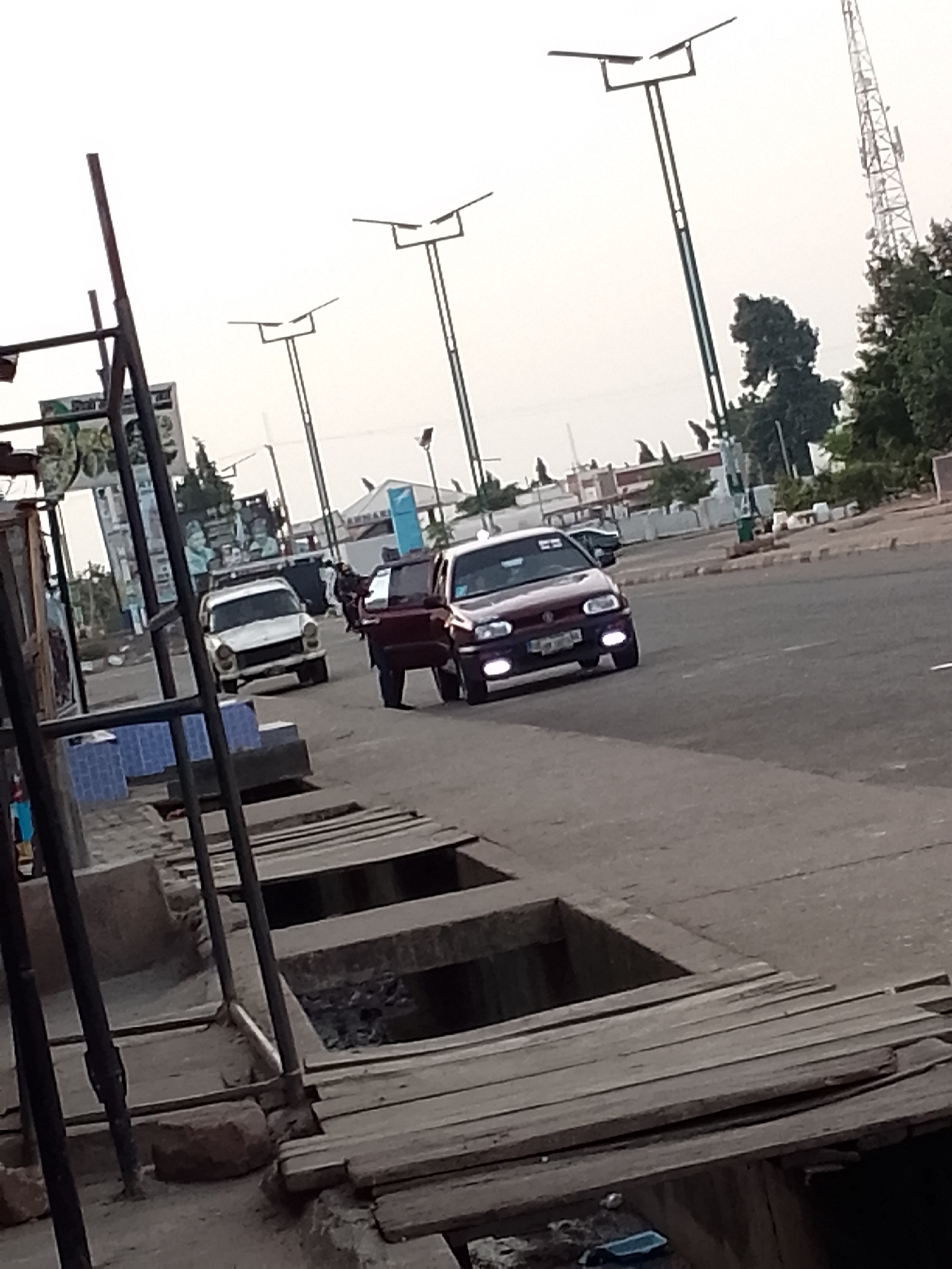 The image depicts a street scene in Pantame, Nigeria. In the foreground, a weathered wooden structure with several open rectangular pits runs along the side of the road. These pits appear to be part of a drainage system, with some showing signs of debris. A narrow paved road occupies the middle ground, with two cars driving on it. The car on the left is a white sedan, while the car on the right is a darker colored hatchback. In the background, streetlights with solar panels are visible, along with trees and buildings. A cellular tower stands prominently to the right. The sky is overcast.