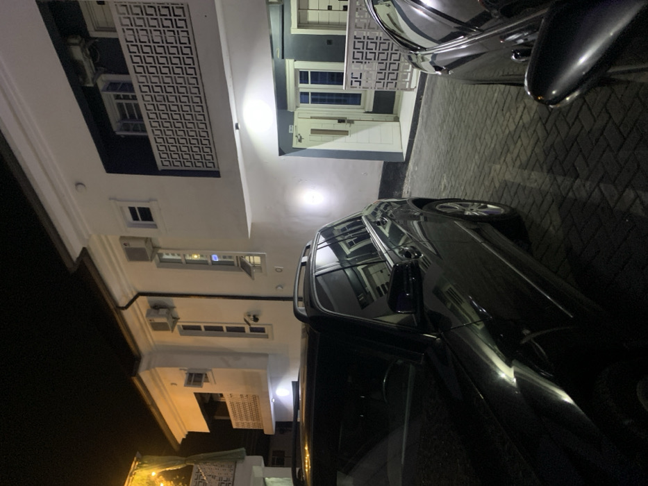 The image depicts an exterior night scene in Iranla, Nigeria, showcasing a multi-story building and two parked vehicles on an interlocking brick pavement. The building features white and dark grey/blue facades, multiple windows, and balconies with decorative white geometric pattern railings. Several wall-mounted air conditioning units are visible on the building's exterior. Artificial lights illuminate the building's facade and the paved ground. On the left, a dark-colored SUV, identifiable by its roof rails, is partially visible. On the right, the front portion of another dark-colored vehicle, likely a sedan, is partially visible. No individuals are present in the scene.