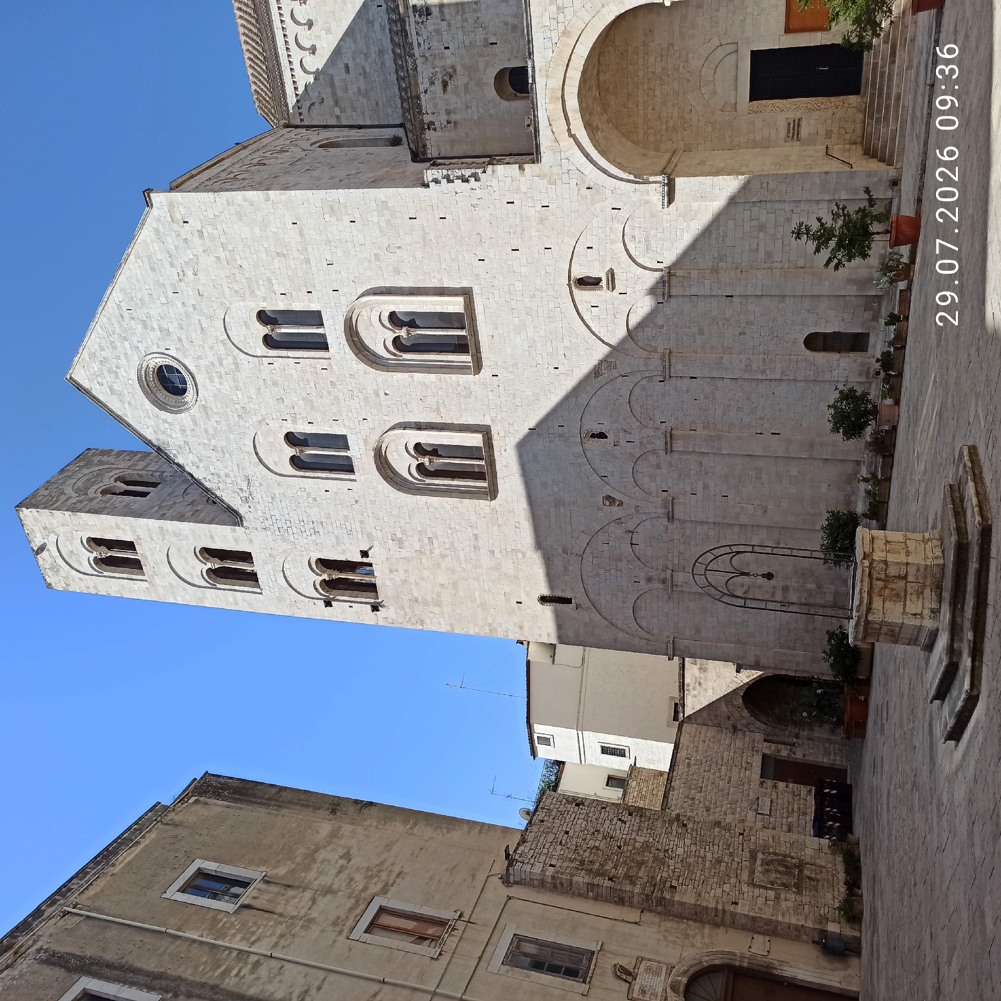 The image depicts an outdoor plaza in front of the Basilica Cattedrale di Maria Santissima Annunziata in Bitonto, Italy. The facade of the basilica, constructed from light-colored stone, dominates the right side of the frame, featuring a tall bell tower with arched windows and arched entrances at its base. The basilica's facade is adorned with multiple arched windows of varying sizes. A circular window is visible on the upper part of the building. The plaza is paved with cobblestones. In the foreground, a stone well with an wrought iron structure above it stands in the center of the plaza. Several potted plants are scattered around the plaza, and there are trash bins visible near a stone wall on the left side of the image. The left side of the image shows the exterior of another building, also constructed of stone, with several windows. The sky is clear and blue. The date and time "29.07.2026 09:36" are superimposed on the lower right corner of the image. No people are visible in the scene.