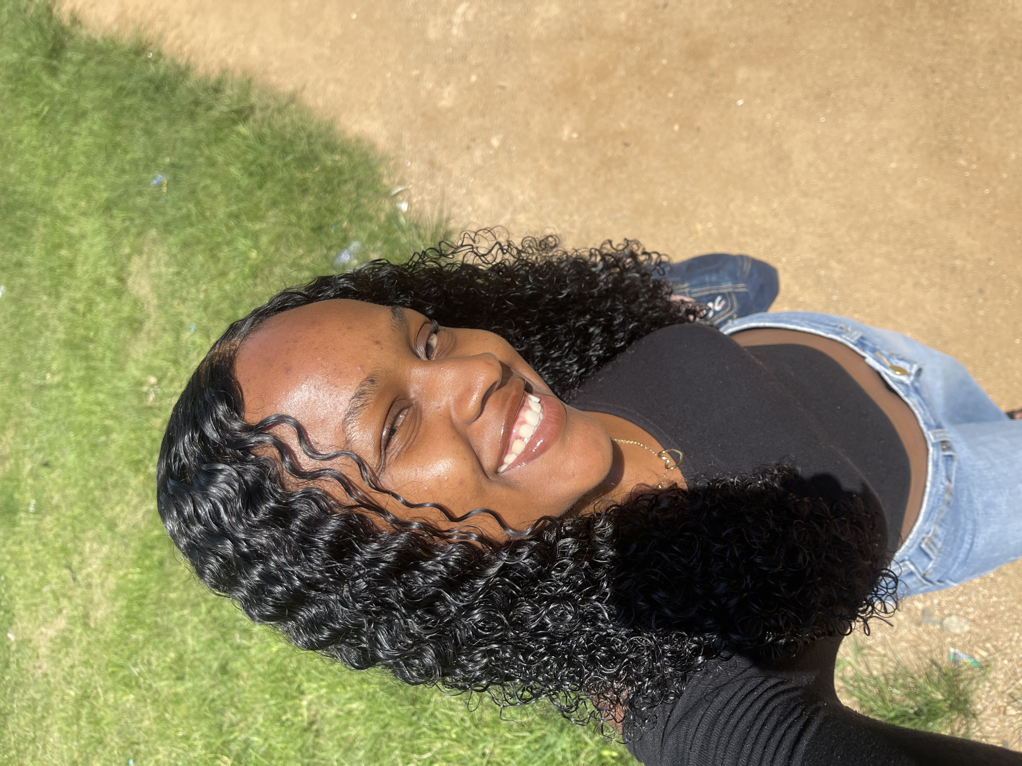 A medium shot from above shows a young Black woman with curly dark hair smiling and winking at the camera. She is wearing a black crop top and light blue jeans. A portion of a dirt path is visible to her right, and a patch of green grass is to her left. The scene is illuminated by direct sunlight, casting shadows. The location appears to be an outdoor area, potentially in Rafin Par, Nigeria.