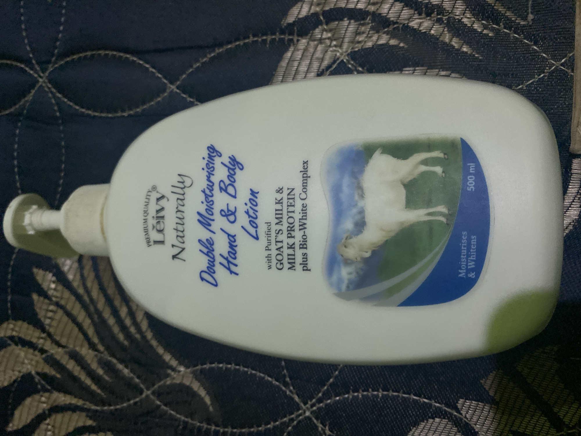 A white pump bottle of Leivy Naturally Double Moisturising Hand & Body Lotion is positioned on a dark, patterned fabric. The bottle features blue text and a picture of a white goat in a pastoral setting. The text indicates the lotion contains purified goat's milk, milk protein, and a Bio-White Complex, and it moisturizes and whitens. The bottle's volume is stated as 500 ml. The fabric appears to be a tablecloth or bedspread with a repeating pattern of stylized leaves or feathers in shades of dark blue and gold, with visible stitching creating lines and curves. The lighting in the image is dim, suggesting an indoor environment. The specific location in Ta', Indonesia, cannot be determined from the visual information provided in the image.