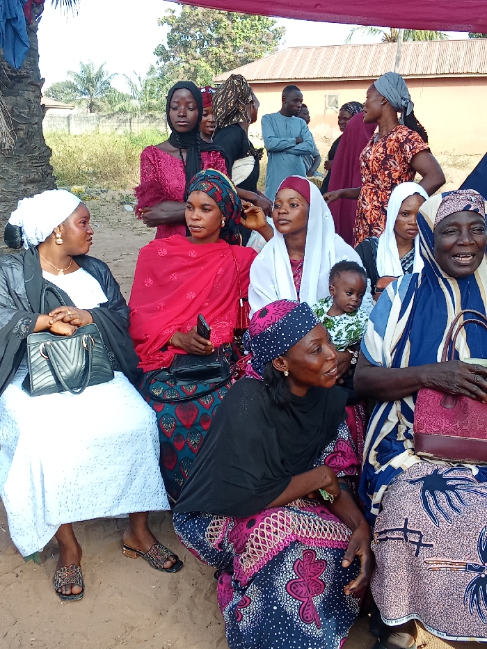 This image captures an outdoor gathering of people, predominantly women, in Doma, Nigeria, under bright daylight conditions. A red canopy or tarp is visible overhead, suggesting a planned event or temporary shelter.
In the foreground, several women are seated or squatting on the dirt ground. On the far left, a woman in a white dress, black outer garment, and white headscarf sits, holding a black handbag, with patterned sandals on her feet. Beside her, a woman in a red headscarf and a dark patterned skirt holds a mobile phone. Next, a woman in a white headscarf cradles a baby dressed in a light green and white patterned outfit. To the right, an older woman in a blue and white striped garment and patterned skirt smiles, holding a pink handbag. Another woman in a black headscarf and a vibrant patterned skirt is seen squatting low in the immediate foreground, looking to the right.
In the midground, a young woman in a black headscarf and a dark red patterned dress stands with her arms crossed, looking downwards. Other individuals, including a few men, are scattered behind the seated group. One man in a light blue tunic stands with his arms crossed. Most women wear head coverings and brightly colored, patterned traditional attire, while the men are also in traditional wear. The expressions on the faces of the subjects vary from attentive and conversational to smiling and pensive.
The background shows a simple setting with dirt ground, sparse vegetation including a prominent palm tree trunk on the left, and basic buildings with corrugated iron roofs under what appears to be a clear sky. No visible text is discernible in the image. The scene conveys a sense of community or family social interaction.