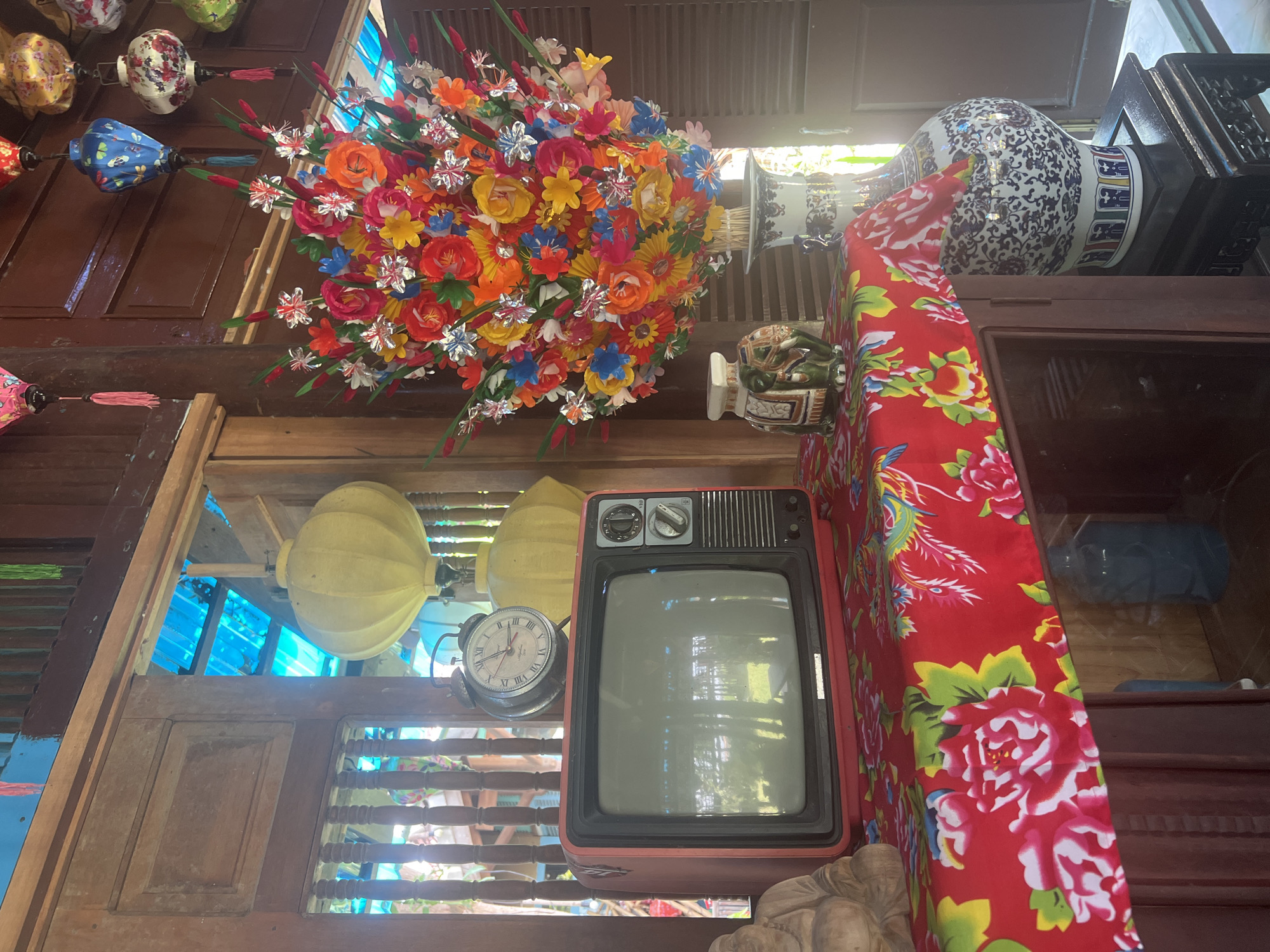 This is a medium shot, eye-level, indoor photograph of a still life arrangement on a wooden cabinet. The scene is brightly lit with natural light, casting some reflections. Dominating the center of the frame is a vintage red-cased television with a blank screen, positioned on a red tablecloth adorned with large floral patterns in pink, white, and green. To the left of the television, a wooden statue of a seated figure, possibly a Buddha, is partially visible. Behind the television, an old-fashioned alarm clock with a Roman numeral face sits on the cabinet. Above the clock, two large, yellow, paper lanterns hang, appearing to be part of a decorative display.
To the right of the television and floral arrangement, a large, colorful, artificial flower bouquet is displayed in a white vase with blue floral patterns. The bouquet consists of numerous vibrant flowers in shades of red, orange, yellow, blue, and pink, with some silver accents. To the left of the flower bouquet and in front of the vase is a small, decorative ceramic elephant figurine with intricate patterns.
The background consists of wooden walls and doors, with a glimpse of blue translucent material, possibly a roof or awning, through an open doorway or window. Several decorative paper lanterns in various colors and patterns, with tassels, are visible hanging from the ceiling or an upper structure. The overall aesthetic suggests a decorative or cultural setting, possibly a shop or a home in Hoi An, Vietnam, known for its traditional lanterns and arts. The photograph captures a collection of decorative items, emphasizing color and texture.