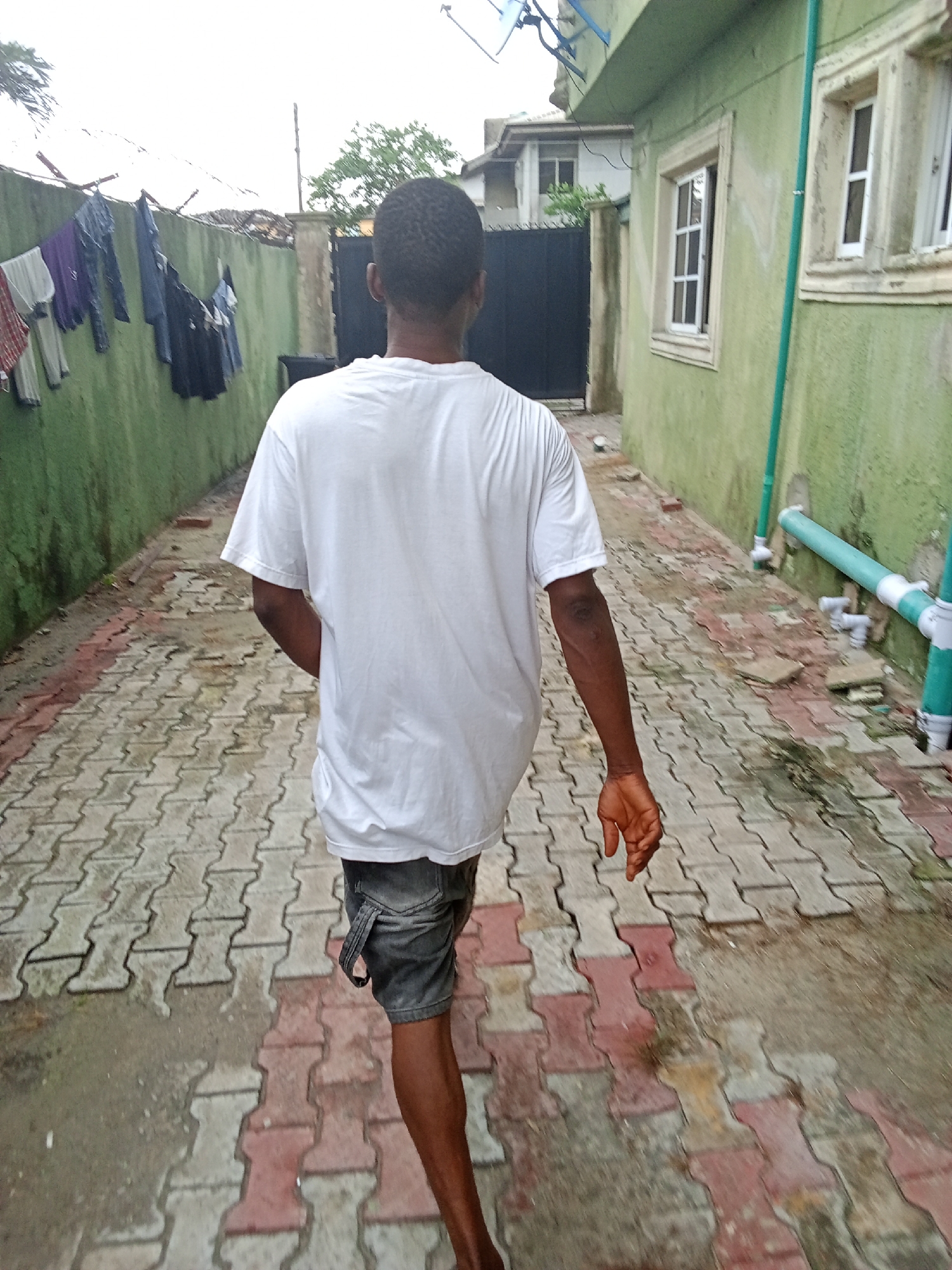 The image depicts a person walking away from the camera down a narrow outdoor pathway in Ado, Nigeria. The individual, a male with dark skin and short dark hair, is seen from behind, wearing a white short-sleeved t-shirt and grey shorts. His left arm is slightly bent, with his hand visible.
The pathway is paved with interlocking concrete blocks in a mix of red and grey, appearing somewhat damp or stained. On the left side of the path, a tall, weathered green wall is topped with barbed wire, and several items of clothing are hung to dry on a line strung across it. To the right, a building with a similarly weathered green facade features two white-framed windows and a series of green and white plumbing pipes running vertically and horizontally along its exterior.
In the background, a dark gate is partially visible, beyond which another building and some trees can be seen under an overcast sky. The diffused lighting suggests a cloudy day, possibly early morning or late afternoon. The scene portrays a everyday outdoor setting, likely within a residential area. No visible text is present in the image.
