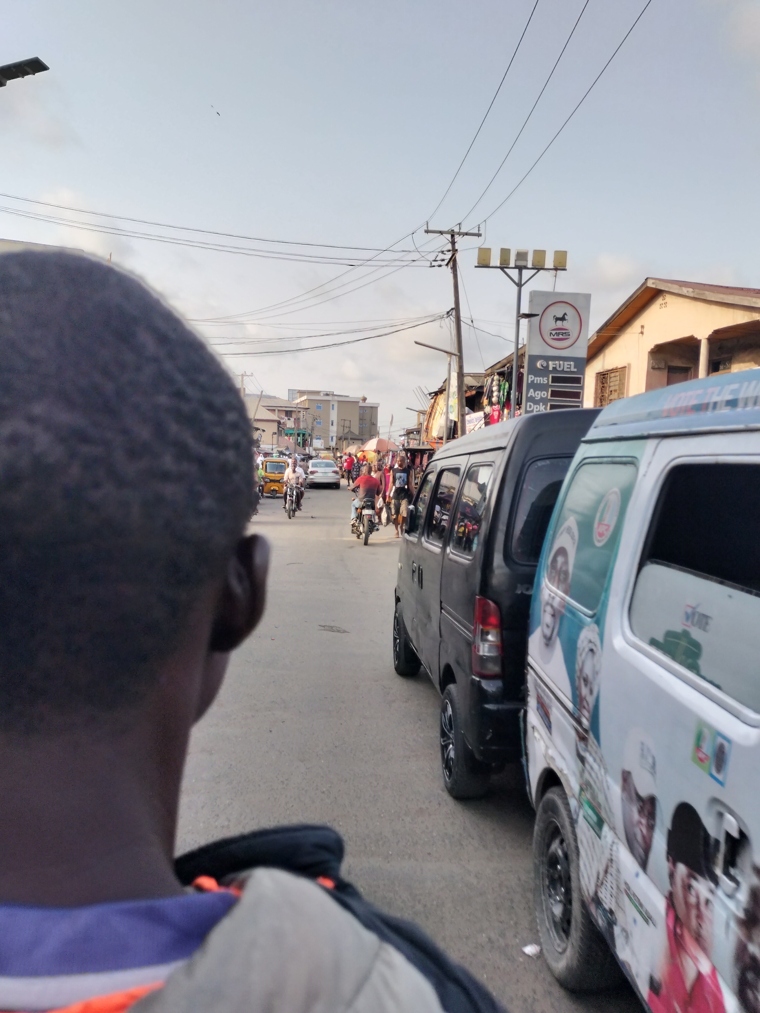 The image presents an urban street scene in Lagos, Nigeria. A partial view of a dark-skinned person's head and shoulder is in the foreground. The street shows vehicular traffic including black and white vans with political campaign images, a yellow tricycle taxi ("Keke Napep"), motorcycles, and cars. People walk along the roadside. A filling station with an MRS Fuel sign is visible. Power lines stretch across the sky. The environment appears to be a busy commercial area with buildings and shops lining the street.