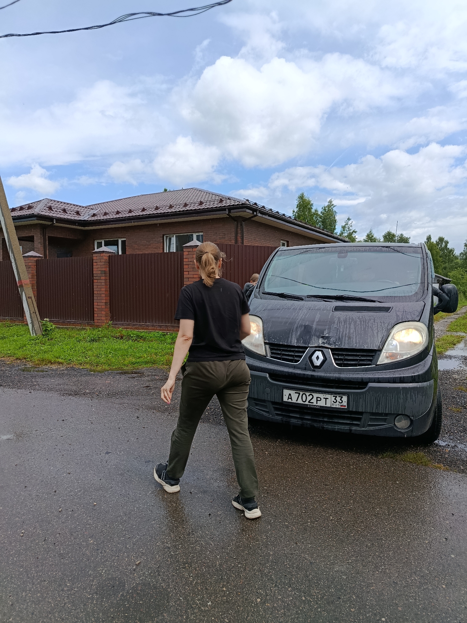 A woman in a black t-shirt and olive-green pants walks away from the camera towards a dark grey Renault Trafic van. She has her hair pulled back in a ponytail. The van's headlights are on, and its windshield and hood are wet, suggesting it has been raining. A license plate is visible on the front of the van, reading "A 702 PT 33 RUS". The scene is set on a wet asphalt road, with a brown brick house and fence in the background. Tall green trees are visible beyond the fence, and the sky is partly cloudy, indicating overcast weather, possibly after a rain shower. A utility pole with concrete base stands to the left of the frame. The overall atmosphere is that of a quiet, residential area in Russia, likely during the daytime.