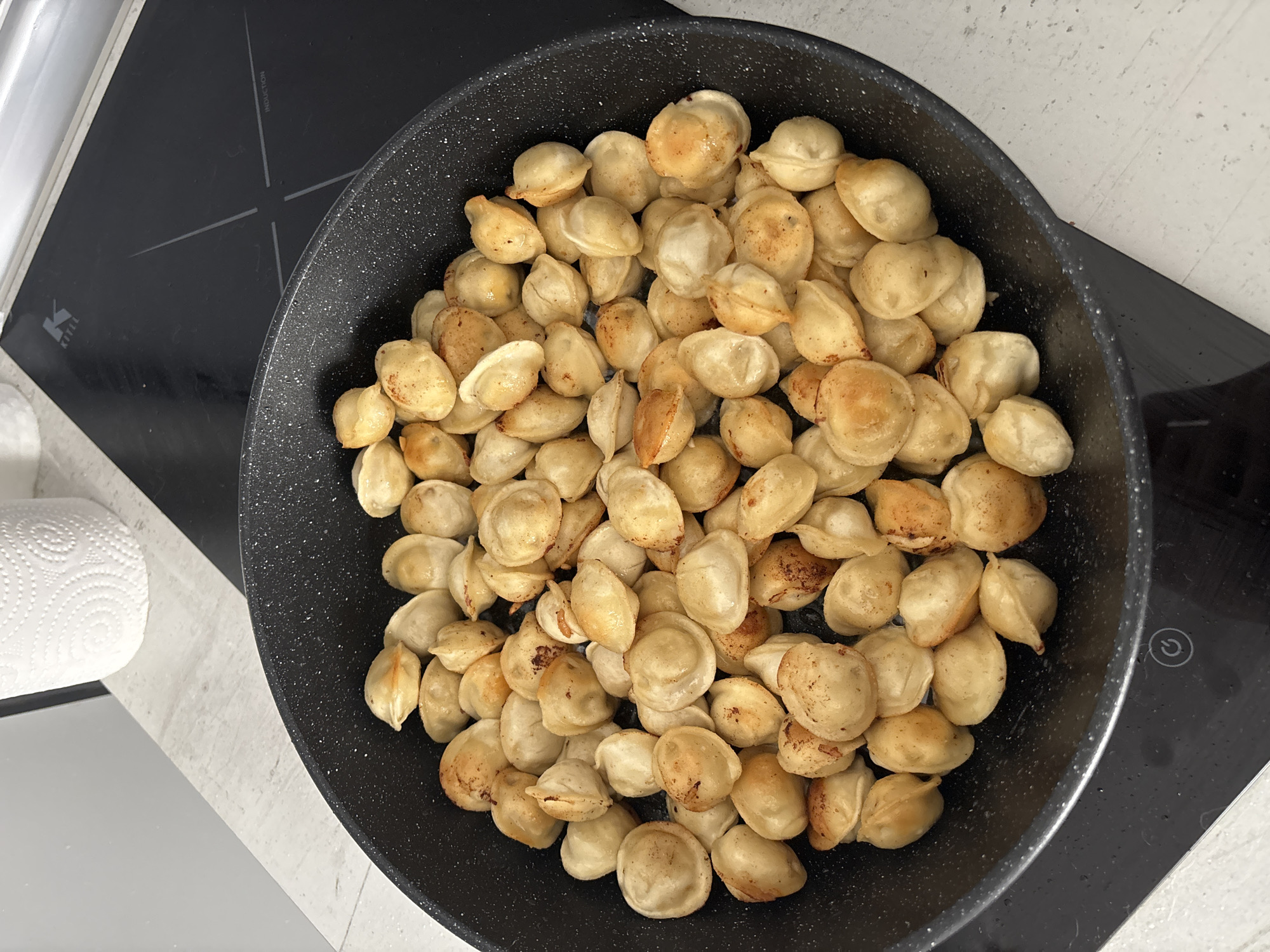 A top-down view shows a black frying pan filled with fried pelmeni (dumplings) resting on a black induction cooktop. To the left of the cooktop is a roll of white paper towels on a light-colored countertop. The scene appears to be indoors, likely in a kitchen. The location is Vsevolozhsk, Russia, based on inference.