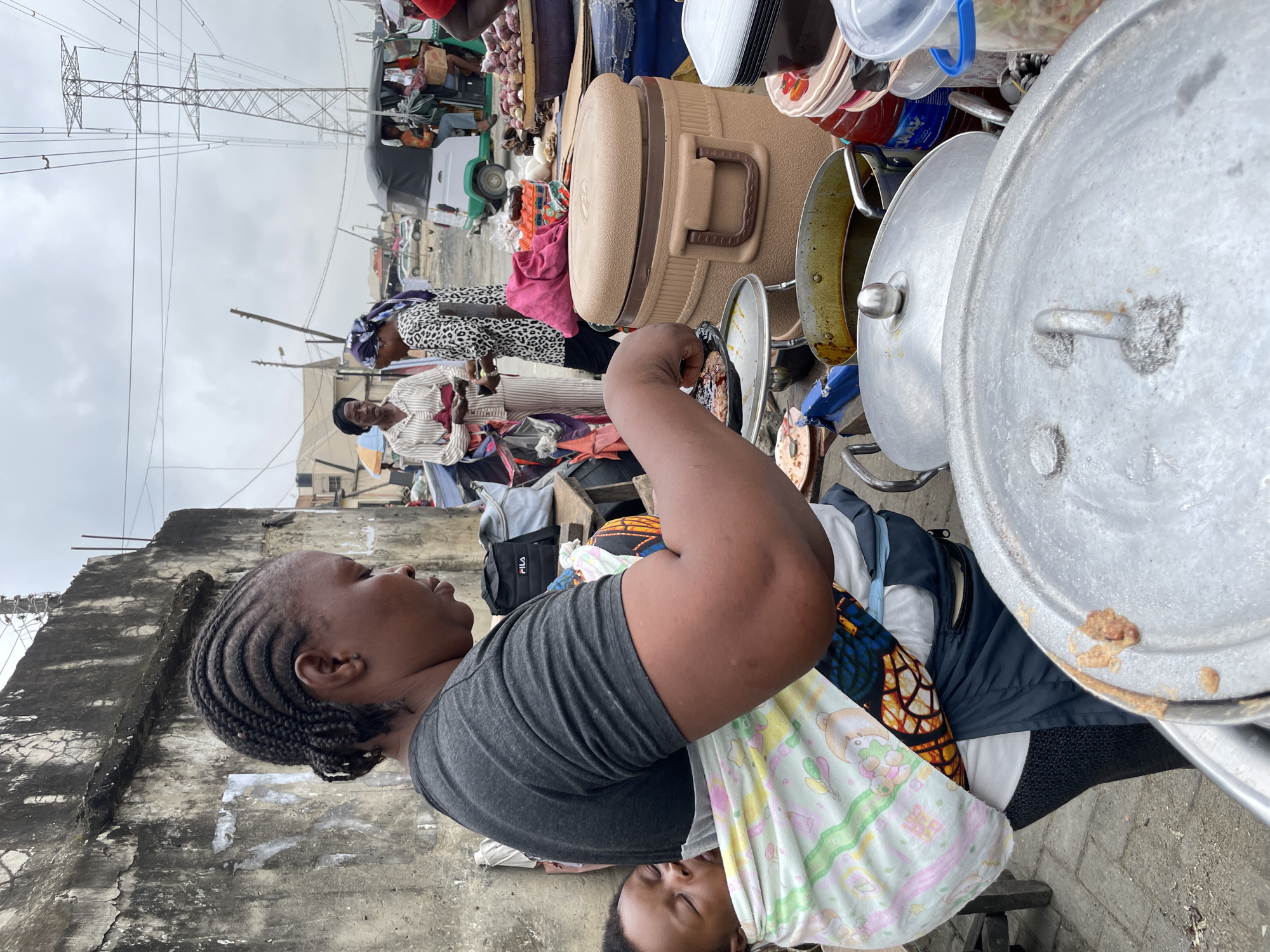 In Ado, Nigeria, a woman is shown from the back carrying a baby wrapped in a colorful cloth sling. She wears a grey t-shirt and dark pants. She is tending to food being cooked in large pots and pans in an outdoor market setting. Behind her, other people are visible, including two women conversing and a person near a three-wheeled vehicle. In the background, there are market stalls, buildings, and a tall electrical pylon, suggesting a busy urban environment. The ground appears to be paved.