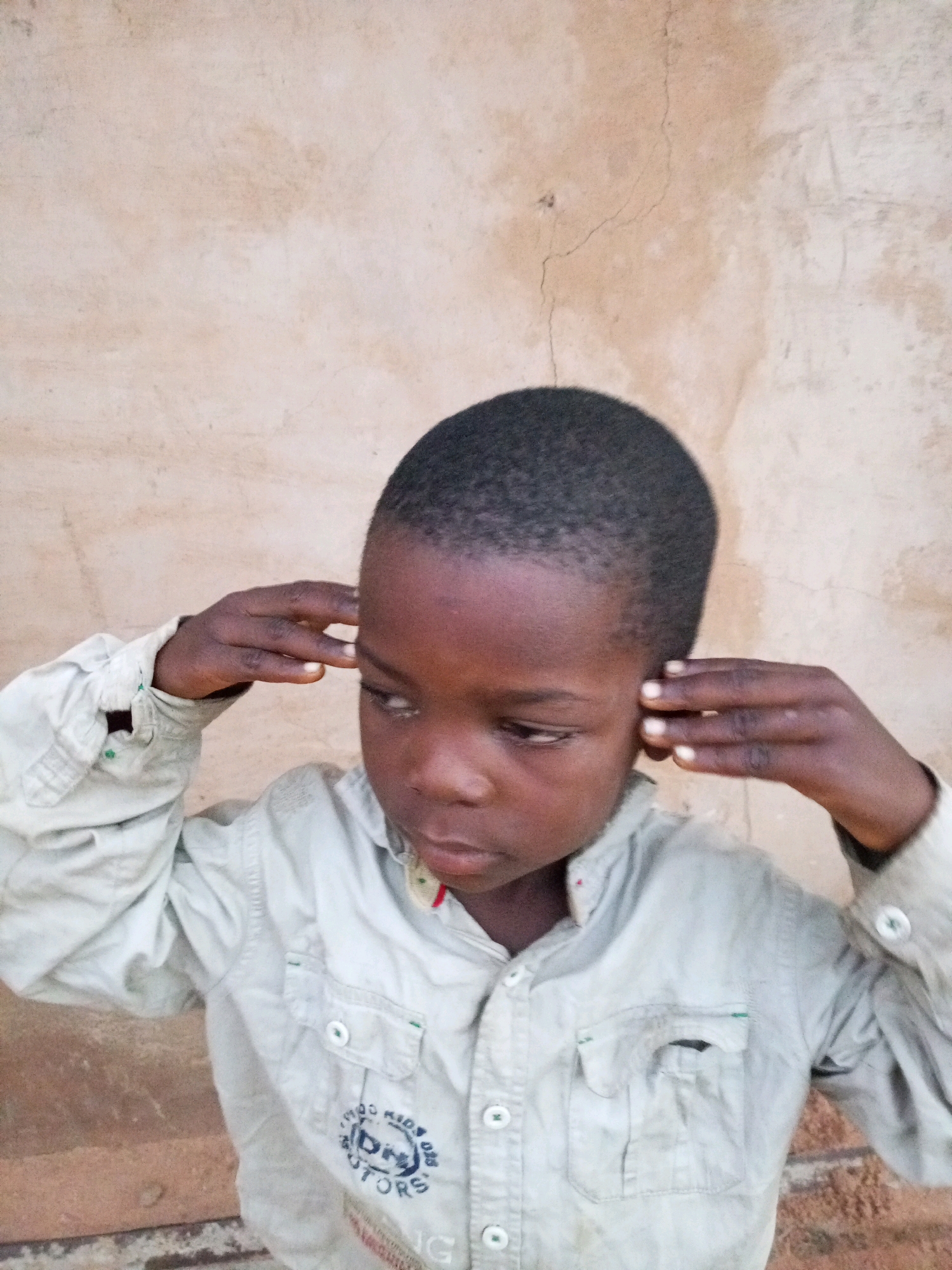 A young Black boy, appearing to be around 5-7 years old, stands in front of a textured, light brown wall that shows some cracks and discoloration. He is wearing a light-colored button-up shirt with two pockets on the chest. The shirt has some green stitching visible on one of the pockets and near the cuffs. The boy's hair is short and dark. He is holding his hands up to his ears, with his fingers touching the sides of his head, as if listening intently or blocking out sound. His gaze is directed downwards and to his right, and his expression is neutral. The lighting suggests it is daytime. The image quality is somewhat grainy, with some overexposure on the wall. The lower part of the image shows a bit of a brick or concrete structure. There is no specific identifiable landmark within the image to pinpoint a location in Tempure, Nigeria.