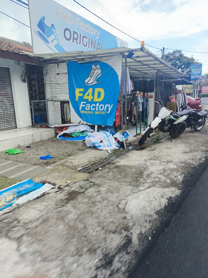 The image shows the exterior of a store called "F4D Factory Original Import", located in Lembang, Indonesia, at latitude -6.822 and longitude 107.61. There is a sign on the storefront with the store name and a blue and yellow logo depicting a sneaker. The sign is above a blue banner displaying the same logo and the words "F4D Factory Original Import". The storefront is mostly obscured by a metal awning covered in a corrugated metal roof. The store has a large window facing the street, and inside it, there are clothes hanging on racks. To the right of the store, there is a white motorcycle parked on the sidewalk. There are several pieces of clothing strewn across the sidewalk in front of the store, along with several blue and white banners. The sidewalk is paved with gray concrete. The street is paved with asphalt. There are no people visible in the image.