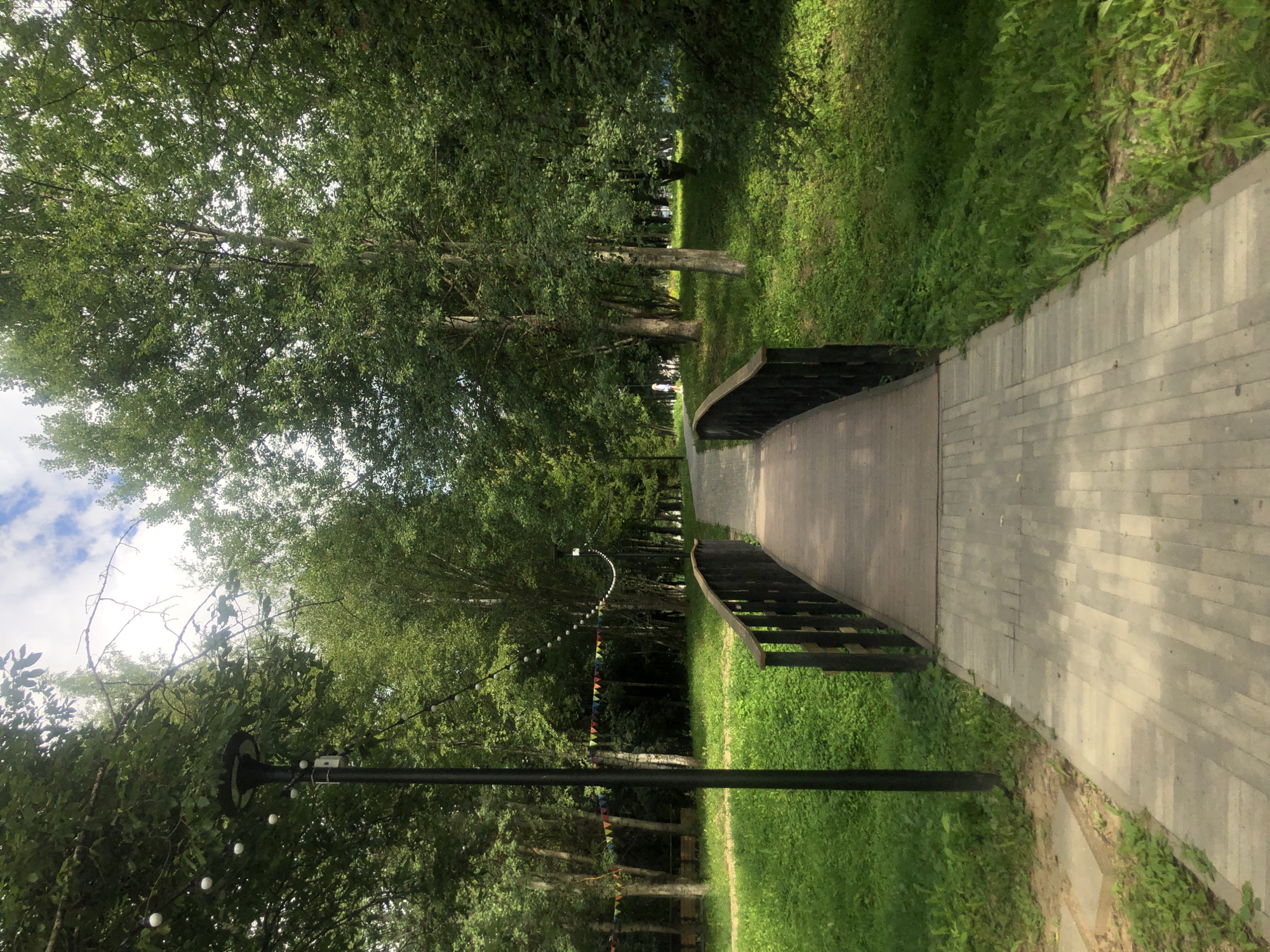 The image depicts a park scene with a paved walkway leading to a wooden bridge. The walkway is made of light-colored rectangular pavers. The bridge has dark wooden railings and a wooden surface. Lush green grass and trees surround the walkway and bridge. In the background, there are more trees, and a bright sky with some clouds is visible. Decorative string lights and small triangular flags are strung between trees on the left side of the walkway. A single street lamp stands on the left side of the walkway, near the foreground. In the distance, on the walkway beyond the bridge, a person in a light-colored outfit can be seen walking away from the camera. The scene appears to be a tranquil park setting, possibly in the city of Tutaev, Russia, given the provided context.