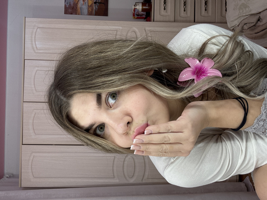 An indoor, close-up portrait shows a female individual, approximately young adult, against a light-colored background in a residential setting in Chaikovsky, Russia. The individual has light brown hair with blonde highlights, styled loosely, and green eyes, wide and looking directly at the camera. Her lips are pursed in a "kiss" gesture, and her right hand, with a French manicure, is brought up to her mouth, partially obscuring her lower face. She wears a light-colored, long-sleeved top, and a grey lace trim is visible on her left forearm. A black elastic hair tie is wrapped around her left wrist. A pink floral hair clip is positioned in her hair on the left side of her head (viewer's right), and a small, silver-colored earring hangs from her left ear. The background features a large, light-colored wooden wardrobe or cabinet with three doors, above which a strip of pink wall is visible. To the right, a section of a lighter wooden dresser or nightstand displays a small item and a pump-bottle dispenser. A small piece of wall art depicting a sunset or sunrise over water with a silhouetted figure is visible further right on the wall.
