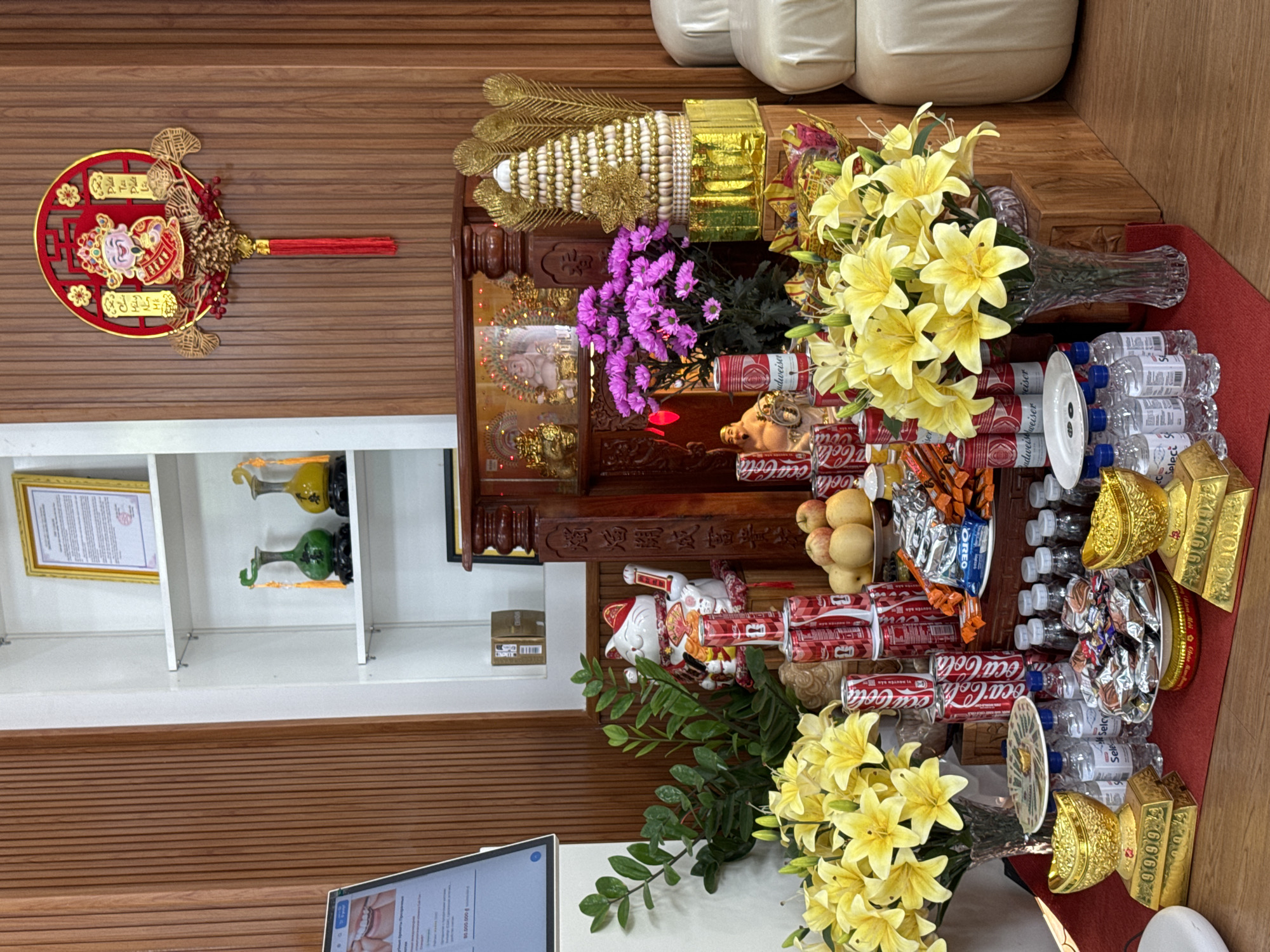This indoor image, taken in Phu Thuy Ward, Vietnam, depicts a vibrant and elaborate display of offerings, likely for a ceremonial or commemorative event such as a traditional celebration, business opening, or ancestral veneration.
The central focus is a wooden altar or display table laden with numerous items. On the table, there are large bouquets of bright yellow lilies and smaller purple chrysanthemums. A golden, tiered ornament with peacock feathers and a decorative golden box stand prominently. Offerings include multiple cans of Coca-Cola and Budweiser, bottles of "Select" brand water, packages of "OREO" cookies, and other wrapped snacks. A bowl of fresh fruit, including apples and pears, is also present. Several symbolic golden ingots, clearly marked "9999," and a white Maneki-neko (waving cat) figurine are arranged amongst the offerings. Small golden statues or figurines are visible on the wooden altar itself.
In the background, a wooden wall paneling provides a warm setting, contrasting with a white shelving unit holding framed documents and decorative gourds or vases. Part of a white upholstered couch or seating is visible in the upper right. On the left, a red circular wall hanging with intricate golden designs and Chinese characters, likely an auspicious phrase ("獨為開成富貴之" can be interpreted as 'May you enjoy wealth and honor'), adorns the wall. In the lower left foreground, a modern computer monitor on a desk displays text, possibly in Vietnamese, and an image of teeth, with "80.000.000 đ" clearly visible, suggesting a monetary value or price. The overall scene suggests a blend of traditional cultural practices with contemporary elements. No people are visible, but the meticulously arranged items imply human involvement in setting up this festive or reverent display.