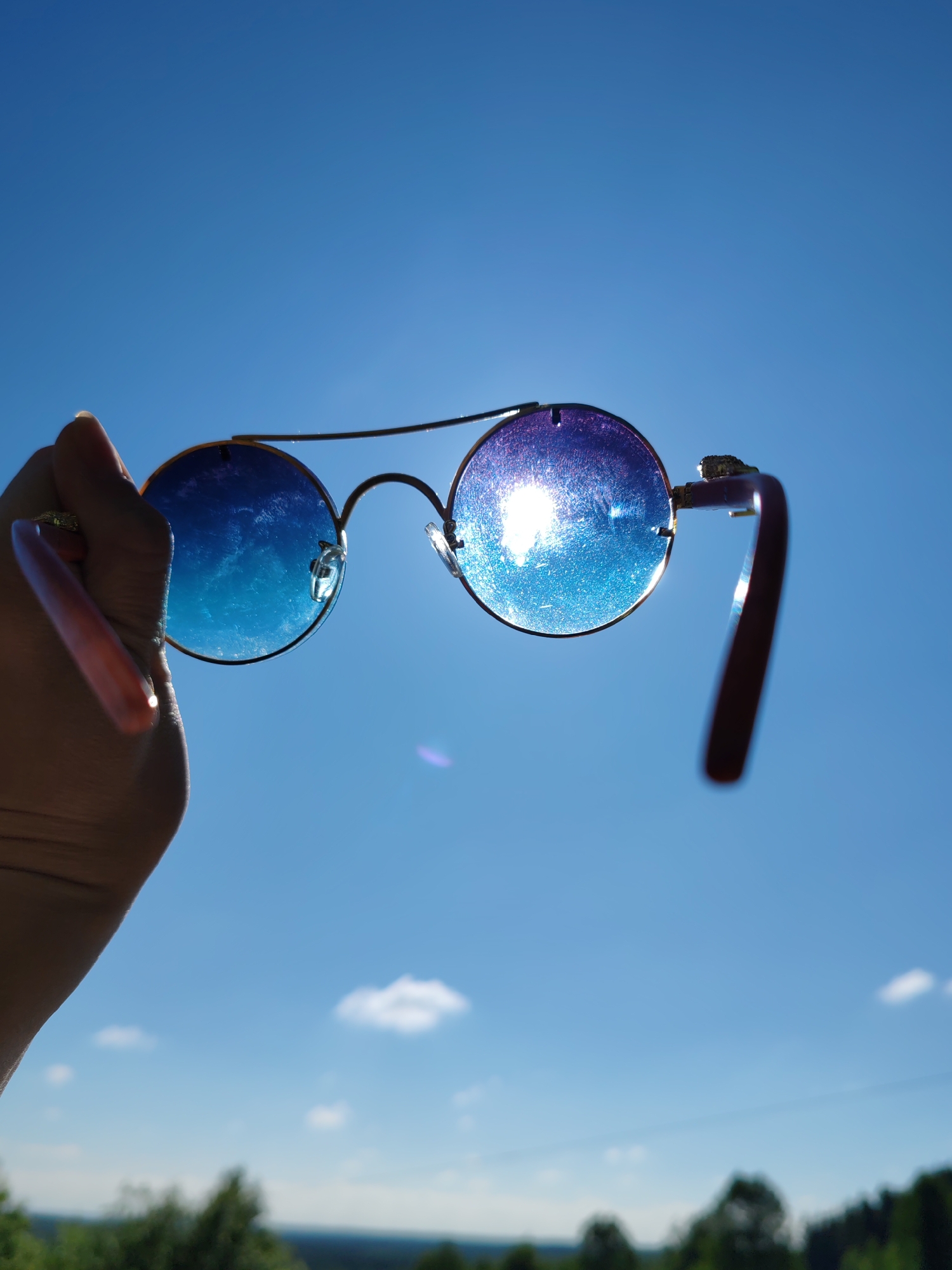 A person's hand, with long fingernails, is holding a pair of round, blue-tinted sunglasses against a bright blue sky. The sunglasses have a gold-colored frame and the lenses reflect the sunlight, creating a dazzling, shimmering effect. The background shows a clear sky with a few wispy clouds, and at the very bottom, a blurry line of trees and distant landscape, hinting at an outdoor, possibly rural or suburban, setting. There is no indication of the city of Чухлэм, Russia.