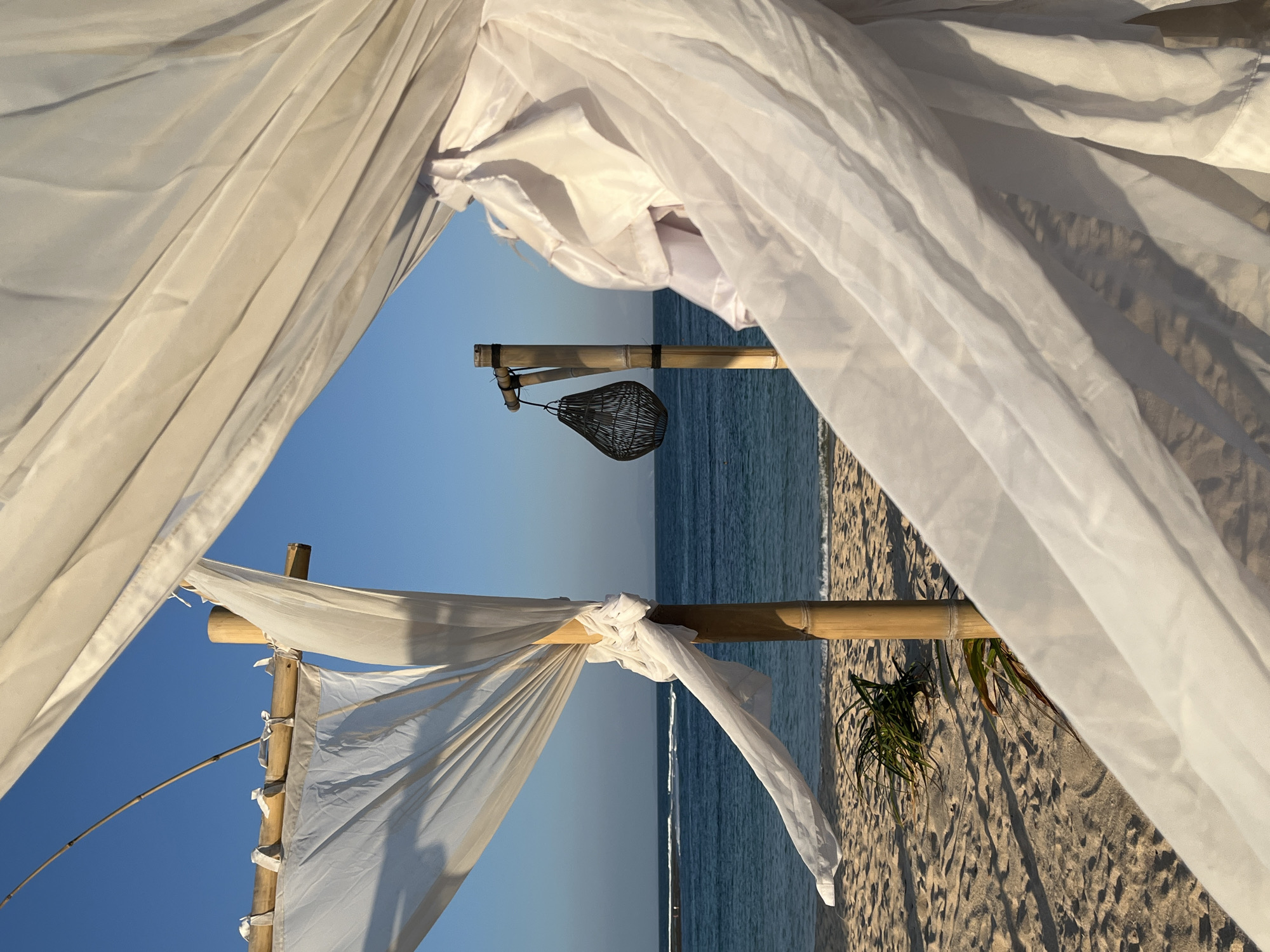 This image depicts a tranquil beach scene at Kuta Selatan, Indonesia. In the foreground, translucent white fabric billows in the breeze, obscuring parts of the frame. Behind the fabric, a bamboo structure supports more white cloth, creating a canopy or tent-like setup. This structure is set up on a sandy beach, with footprints visible in the sand. To the left, a small cluster of green foliage grows. In the middle ground, the turquoise ocean meets the horizon under a clear blue sky. On the right side of the middle ground, a bamboo pole with a dark, woven lantern hanging from it stands. The overall atmosphere is serene and idyllic.