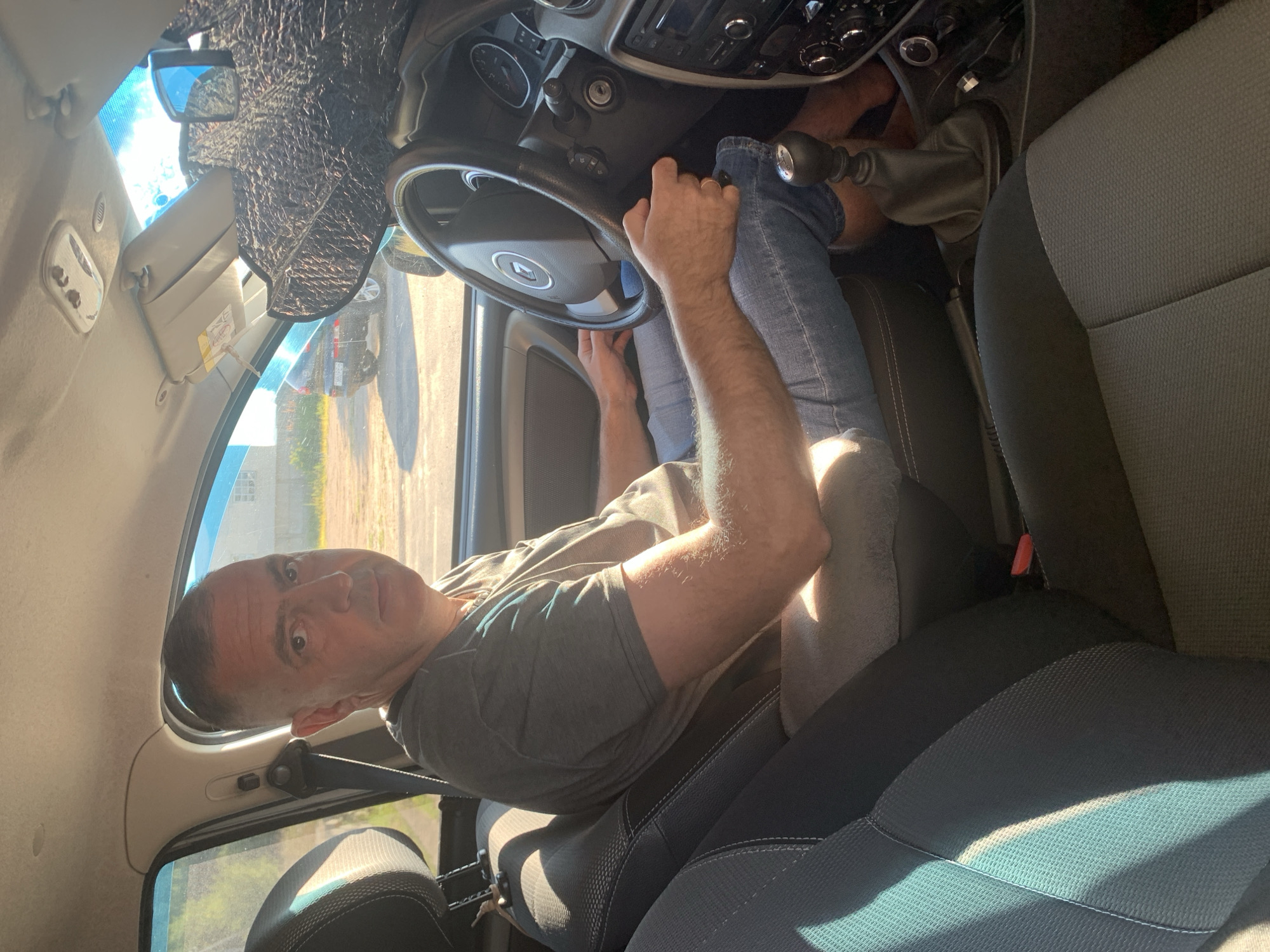 A man is sitting in the driver's seat of a car. He is wearing a grey t-shirt and blue jeans. The car's interior is visible, including the steering wheel, dashboard, and seats. The sun is shining through the windshield, casting shadows on the interior. In the background, another car is parked outside, and a building is visible. The scene appears to be in a rural or suburban area, possibly near the city of Капыльскі сельскі Савет, Belarus.