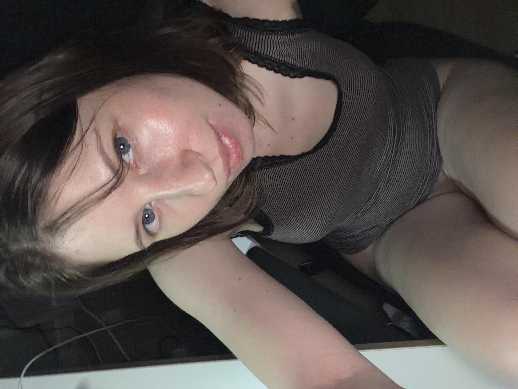 A woman with dark brown hair and blue eyes is lying down, looking at the camera. She is wearing a dark grey and white striped top with a black lace trim around the neckline. The scene appears to be indoors, possibly a room. The lighting suggests it could be nighttime or a dimly lit indoor space. The woman's expression is neutral. Her body is oriented sideways, with her head tilted. The background is dark and out of focus, with some cables visible on a surface in the foreground. No text is visible in the image. The location is specified as Dongducheon-si, South Korea.