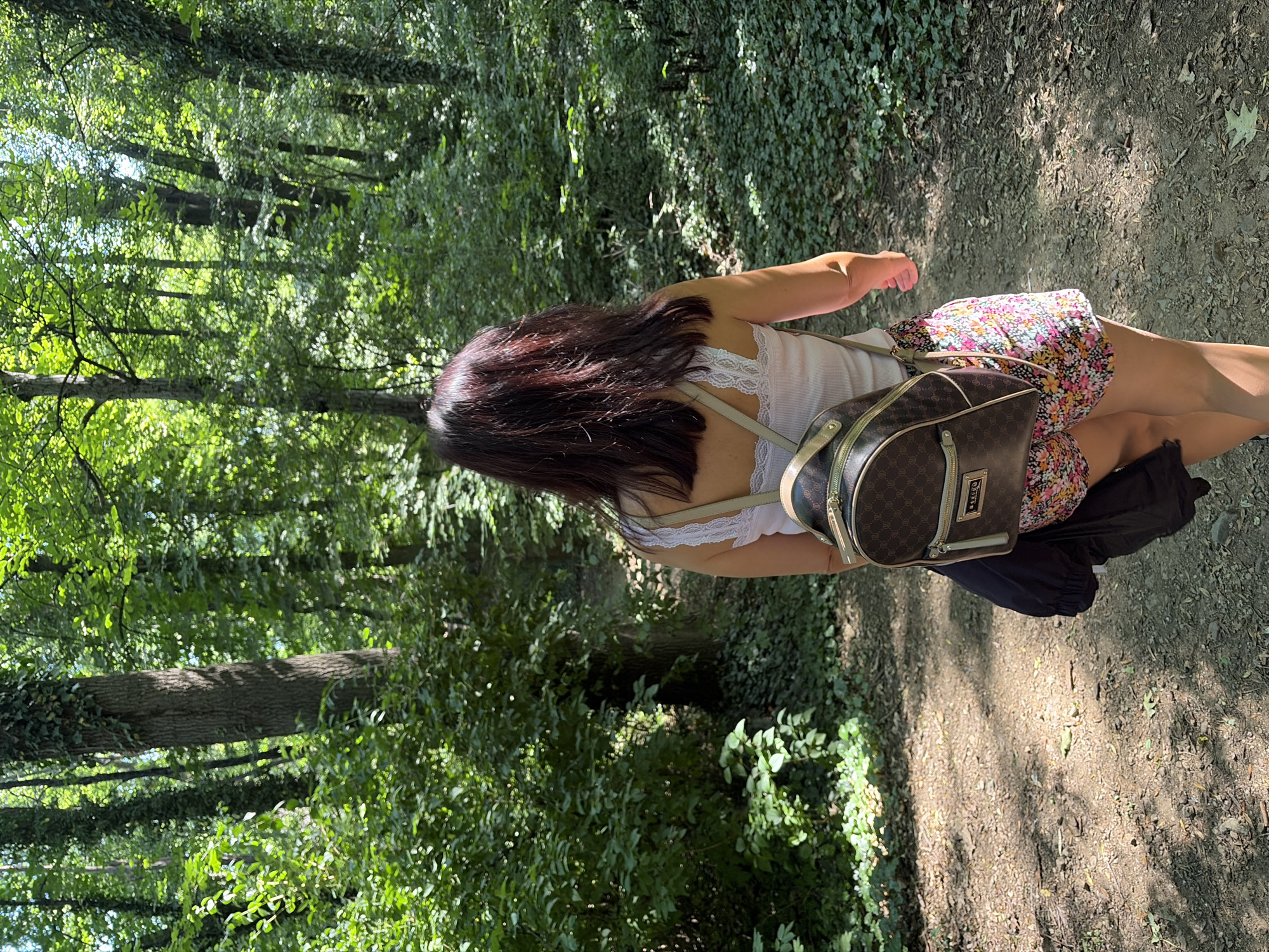 A woman with dark hair walks away from the camera on a dirt path in a sun-dappled forest. She is wearing a white tank top with lace trim, floral print shorts, and a brown backpack with a monogrammed pattern. A black jacket is slung over her left arm. The forest floor is covered with green ivy and fallen leaves. The trees are tall with lush green foliage, indicating a warm-weather season. The scene is likely a park or natural area within Sofia, Bulgaria, given the dense woodland environment and the presence of a well-maintained walking path.