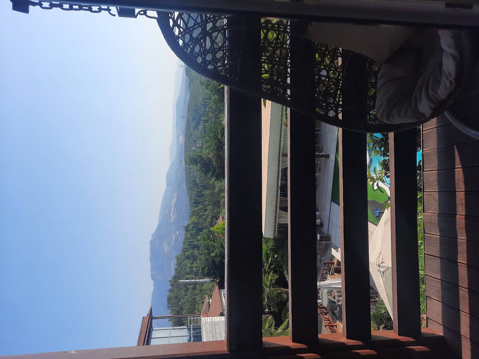 An outdoor balcony overlooks a landscape of rolling hills covered in dense evergreen trees and distant mountains under a clear blue sky. In the foreground, a dark wicker hanging chair with a light-colored cushion is partially visible on the right. The balcony floor is made of wooden planks. Below the balcony, several structures and outdoor amenities are visible, including umbrellas, lounge chairs, a swimming pool, and parts of buildings. The scene appears to be a resort or a home with a scenic view. The location is unknown, Ukraine.