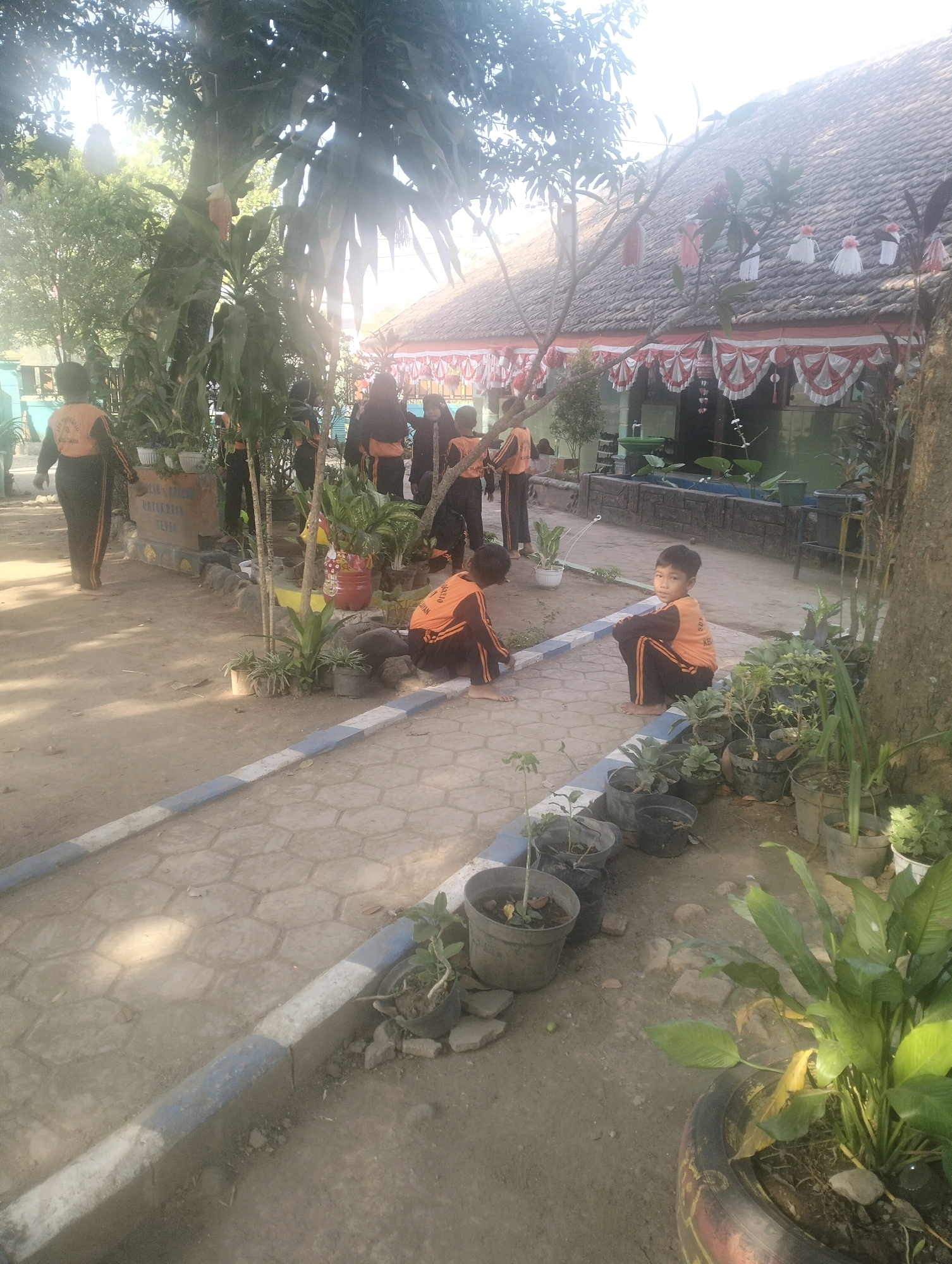 This is a medium-shot, eye-level, outdoor photograph taken in Blabakan, Indonesia. The scene depicts a group of children, likely students, gathered in what appears to be a schoolyard. Several children are wearing orange and black uniforms. In the foreground, two boys are seated on the ground. One boy, facing the camera, is wearing an orange long-sleeved shirt and black pants and is crouched down. Another boy, to his left and partially out of frame, is also wearing similar attire and is seated on the ground.
In the background, a building with a tiled roof is visible, adorned with red and white decorative banners, suggesting a festive occasion. There are also trees and various potted plants scattered around the area, contributing to a natural, somewhat rustic environment. The ground is a mix of dirt and a paved pathway with a hexagonal pattern. The lighting suggests it is daytime, with sunlight filtering through the trees. Several other children in similar uniforms are standing in small groups in the mid-ground, some appearing to be engaged in activities or conversation. The overall atmosphere is that of an informal gathering or activity occurring at an educational institution in a tropical setting.