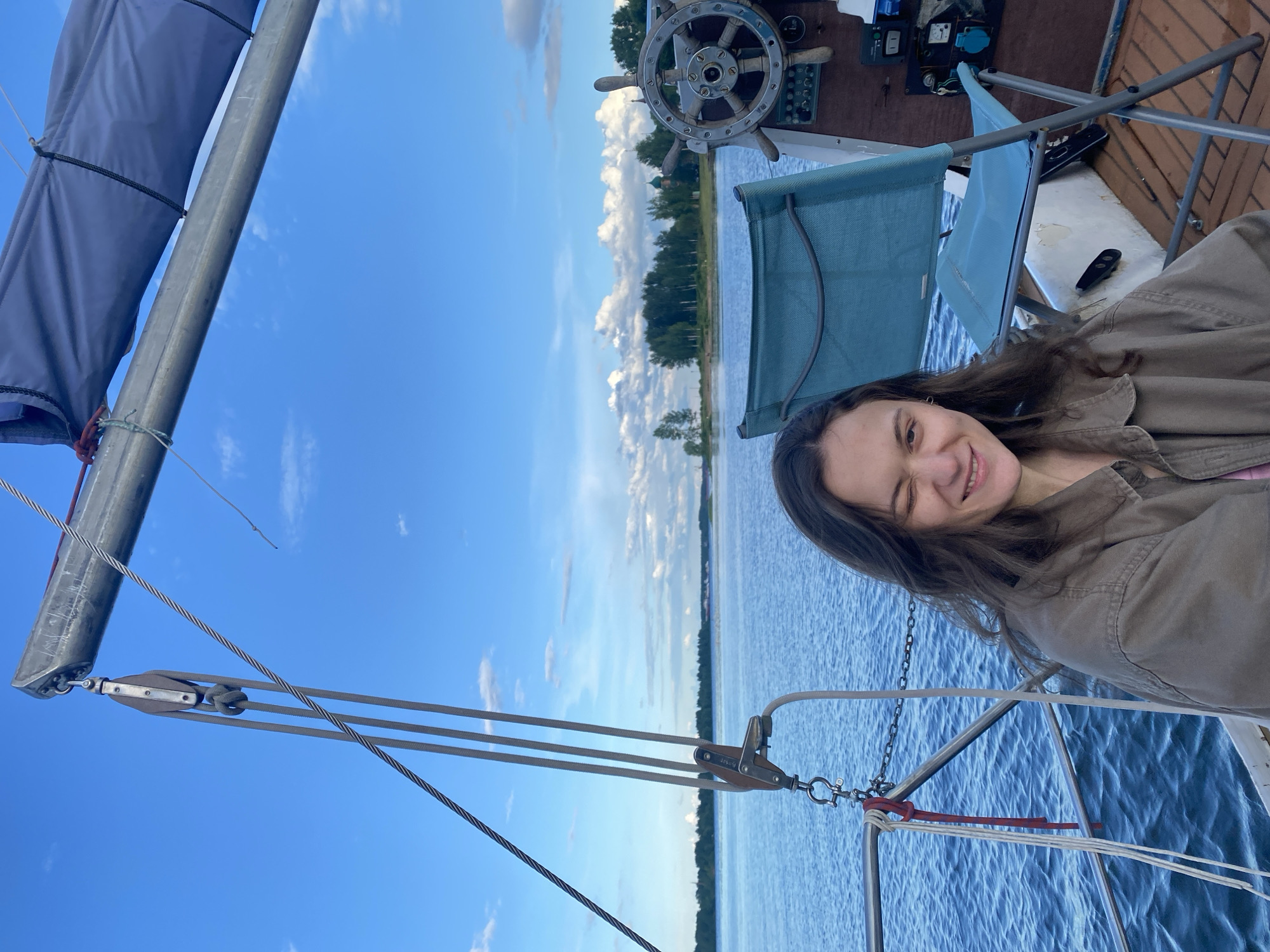 A woman with long, dark, wavy hair winks at the camera while sitting on a sailboat on a body of water, likely a river or lake. She is wearing a light brown jacket over a pink shirt. The sailboat has a large sail deployed and is equipped with a ship's wheel, various ropes, and a blue folding chair. In the background, a tree-lined shore is visible, with some buildings and clouds in the sky. The overall scene suggests a leisurely day on the water.