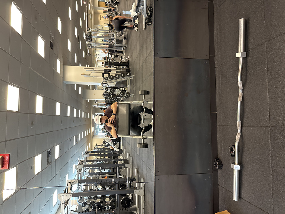 The image shows the interior of a large, well-lit fitness center or gym, located in Boston, United States. The primary subject is a male individual, visible as a reflection in a prominent wall-mounted mirror, seated on a black preacher curl bench. He wears a dark short-sleeved shirt, dark pants, white athletic shoes, black over-ear headphones, and a white baseball cap featuring a red logo (consistent with Callaway Golf branding). He holds a black smartphone in both hands, angled as if capturing a mirror selfie.
In the foreground, on the black rubber mat flooring, lies an EZ curl bar with two black barbell collars positioned nearby. The gym's ceiling is light-colored and features numerous rectangular fluorescent light panels, with a red fire alarm box mounted overhead. The floor throughout the facility is uniformly covered with black rubber mats.
The mirrored reflection reveals an expansive gym space equipped with various weightlifting and exercise machines, including multiple dumbbell racks, power cages, cable machines, and other resistance training apparatus. Several other individuals are visible in the background, exercising. Specifically, a woman in dark athletic wear is observed bending over near a weight bench on the right side of the reflection, appearing to interact with dumbbells or adjust equipment. The environment is modern, organized, and typical of a commercial fitness establishment.