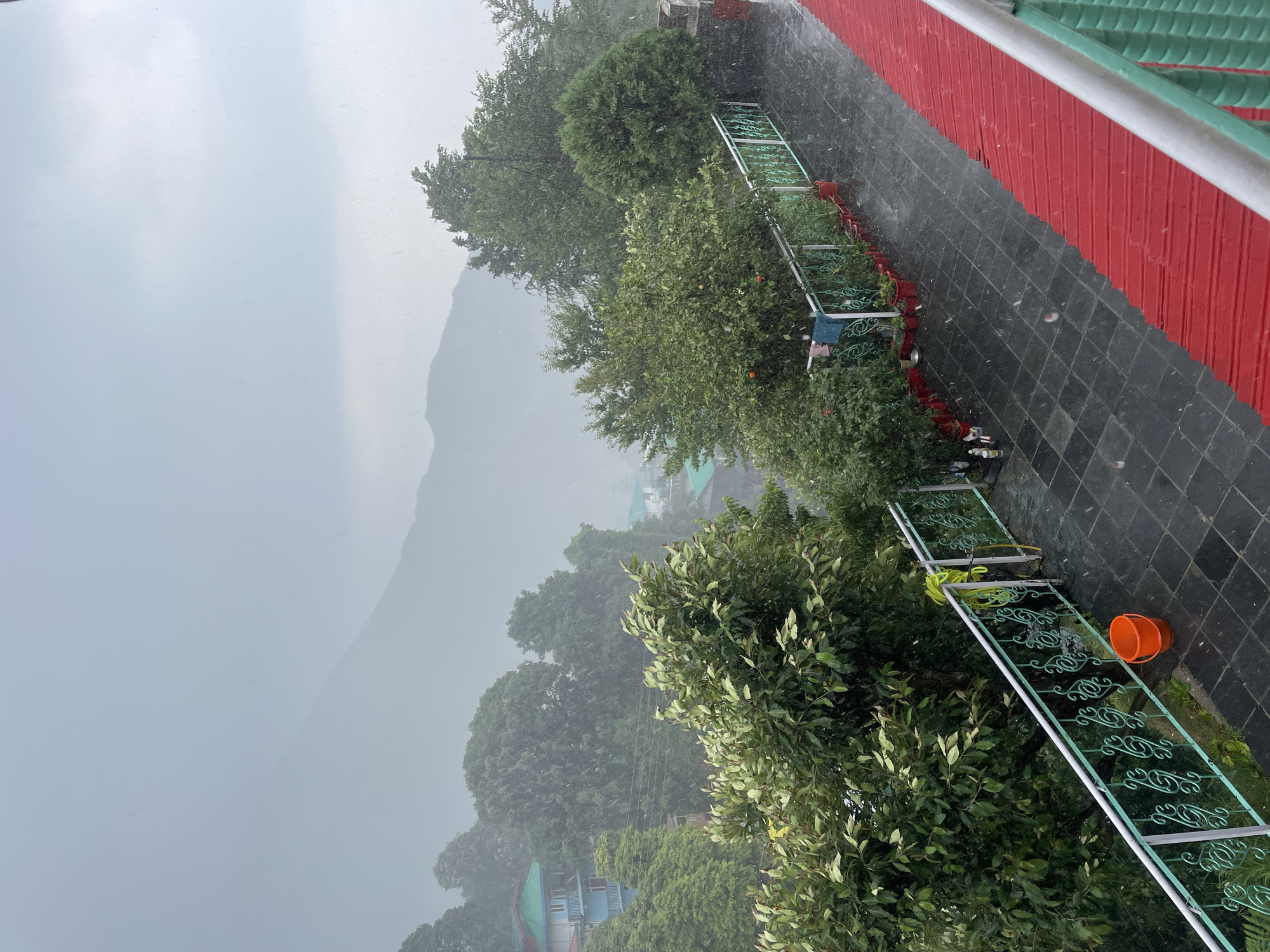 This image, captured in Bhagsu, India, presents a view of a mountainous landscape during a period of active precipitation, likely rain or light hail. The scene is observed from an elevated perspective, possibly a balcony or terrace.
In the immediate foreground, a dark-tiled walkway or patio is visible. To the right, a striking red wall with vertical corrugated or ribbed panels rises, topped by a segment of a green roof. Along the left edge of the tiled area, a decorative green metal railing featuring ornate scrollwork extends horizontally. Beyond this railing, a series of lush green bushes and plants in red pots are arranged. An orange plastic bucket and a coiled yellow hose are also noticeable on the tiled surface.
The midground is densely covered with vibrant green trees and vegetation, some appearing to be fruit trees with small orange-colored fruits. Nestled within this verdant expanse, several buildings with distinctive green roofs can be seen, indicating a settlement in the hilly terrain.
The background features majestic mountains, their contours softened and partially obscured by a thick, grey mist or haze, intensified by the ongoing precipitation. The sky is uniformly overcast and grey, suggesting daylight hours. Numerous white specks and streaks are scattered throughout the image, especially visible against darker elements, confirming the presence of rain or possibly light hail. The overall atmosphere conveyed is one of tranquility and dampness, characteristic of a mountain environment during inclement weather. There are no people or visible text in the image.