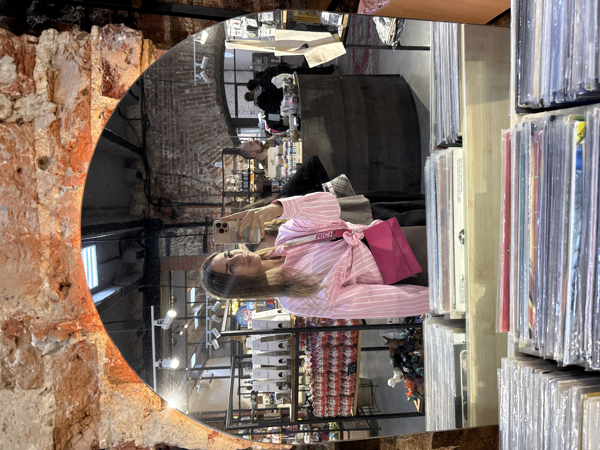 The image shows a woman taking a mirror selfie in what appears to be a record store, potentially in Saint Petersburg, Russia, given the context of the prompt. The woman, with long brown hair, is wearing a pink and white striped shirt, tied at the waist, and a dark skirt. She is holding a smartphone in her right hand, taking the picture. A bright pink crossbody bag is slung across her chest.
In the reflection of the mirror, the store's interior is visible. Shelves are stocked with various items, including what look like boxes of chocolate eggs and other packaged goods. In the background, there are displays of records. A counter area is visible with people interacting, including a man with a beard speaking to someone behind the counter. The architecture features exposed brick walls and arched ceilings, suggesting an older building, common in historic cities like Saint Petersburg. The lighting appears to be from track lights on the ceiling.