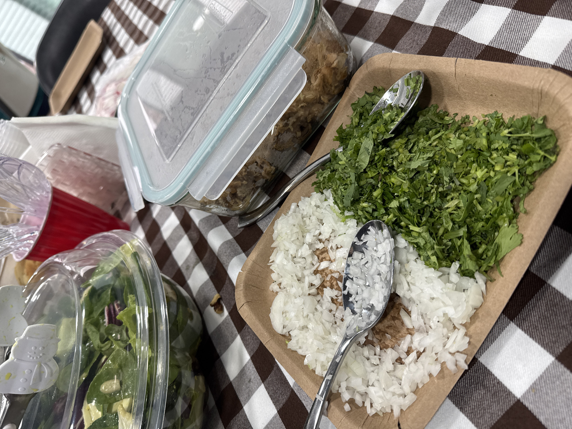The image shows a tabletop scene featuring food items. A brown and white checkered tablecloth serves as the backdrop. In the foreground, there's a clear plastic container filled with a green salad, partially obscured. To the right, a brown paper tray holds chopped cilantro and onions, each with a silver spoon placed on top. Further back, a rectangular clear glass container with a light blue lid holds a brownish-colored food. The location of the photograph is San Bernardino Tlaxcalancingo, Mexico.