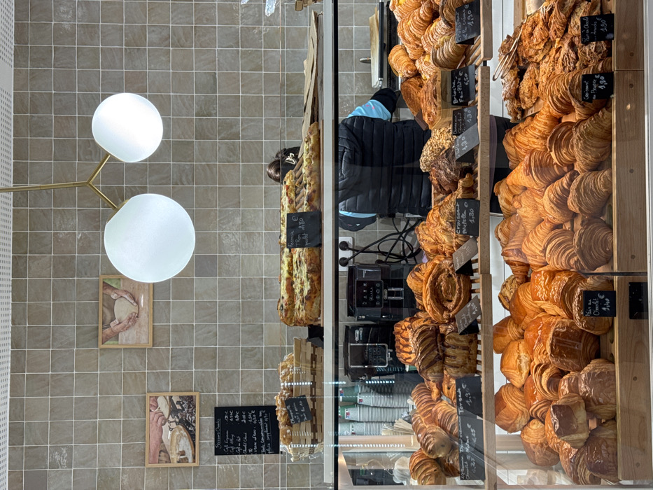 The scene depicts the interior of a bakery or pâtisserie in Asnières-sur-Seine, France. A display counter is filled with a variety of freshly baked goods.
The background wall features light grey-brown square tiles. Two framed pictures adorn the wall, each showing hands working with dough, reinforcing the bakery theme. A modern light fixture with a gold-toned branched frame holds two large spherical white glass globes, providing illumination.
On the upper counter level, two large rectangular savory tarts are visible, one with a small chalkboard label indicating "Focaccia TRom 4,80 €". A wicker basket holds smaller pastries to the left. A black menu board lists various coffee items and prices in French, such as "Café Expresso 1,90 €" and "Chocolat Chaud 3,50 €".
Below the upper counter, behind a glass display, numerous viennoiseries and pastries are arranged on multiple wooden shelves. These include croissants, pains au chocolat, pains aux raisins, and other spiral and layered pastries. Many items have small black chalkboard labels indicating their names and prices in Euros (e.g., "Croissant 1,30 €", "Pain au Chocolat 1,40 €", "Torsade 2,30 €").
Behind the display case, a person is partially visible, facing away from the camera. They wear a dark blue quilted vest over a lighter blue long-sleeved shirt. Their dark hair is pulled back with a light-colored clip. Two black coffee machines (Delonghi brand) and stacks of disposable cups are situated on the counter behind the display, along with electrical outlets and wires. A digital scale is also present on the counter to the right.