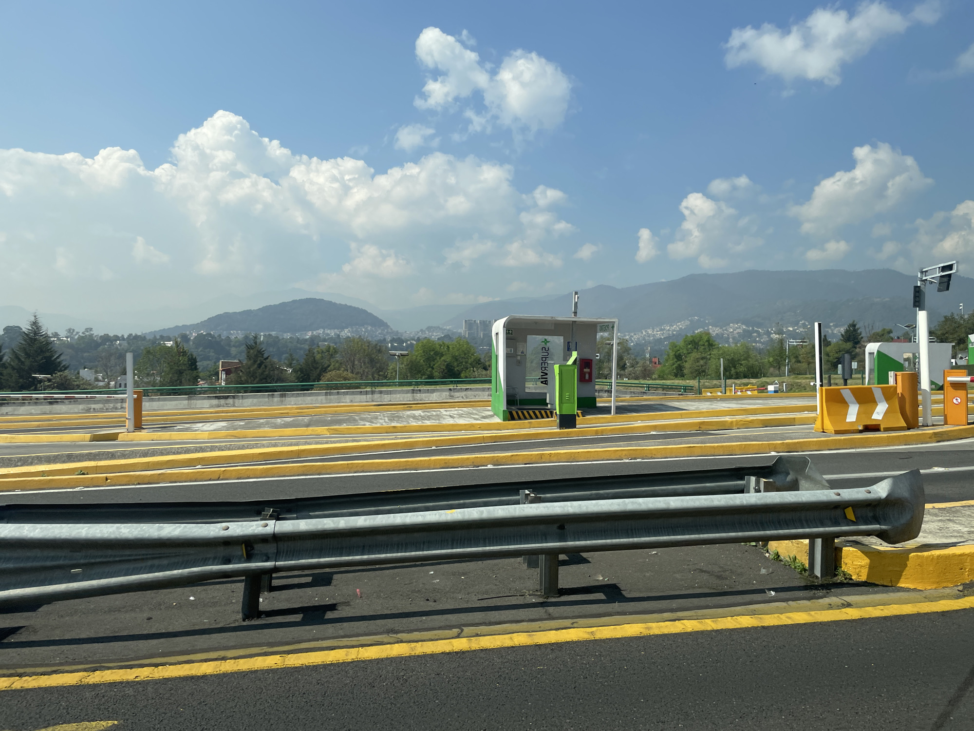 This daytime photograph captures a multi-lane toll plaza or highway entrance, likely situated in Mexico City, against a backdrop of rolling hills and mountains. In the foreground, a dark asphalt road is visible, marked with bright yellow lines, and a sturdy metal guardrail runs horizontally across the lower half of the image.
Beyond the guardrail, several distinct vehicle lanes are visible, separated by prominent bright yellow concrete barriers and curbs. A notable green and white structure, which appears to be a toll booth or automated payment kiosk, stands in the mid-ground. The word "VIA VERDE" is clearly visible on this structure, both vertically on its side and reflected or in reverse horizontally on its front. Another smaller, orange and yellow payment terminal or kiosk is positioned further to the right. White barriers with yellow chevron arrows direct traffic flow. Overhead lighting or signage structures are also present.
The background features an expansive landscape of green, tree-covered hills and mountains stretching to the horizon. Portions of urban development, including houses and buildings, are visible nestled among the trees on the lower slopes. The sky above is a clear, vibrant blue, dotted with numerous fluffy white cumulus clouds.
The scene is brightly lit by natural sunlight, suggesting midday or early afternoon, with distinct shadows cast by the guardrail and structures. The image presents a functional transportation infrastructure set against a scenic natural and semi-urban environment. No people or moving vehicles are visible, emphasizing the structural elements of the toll area.