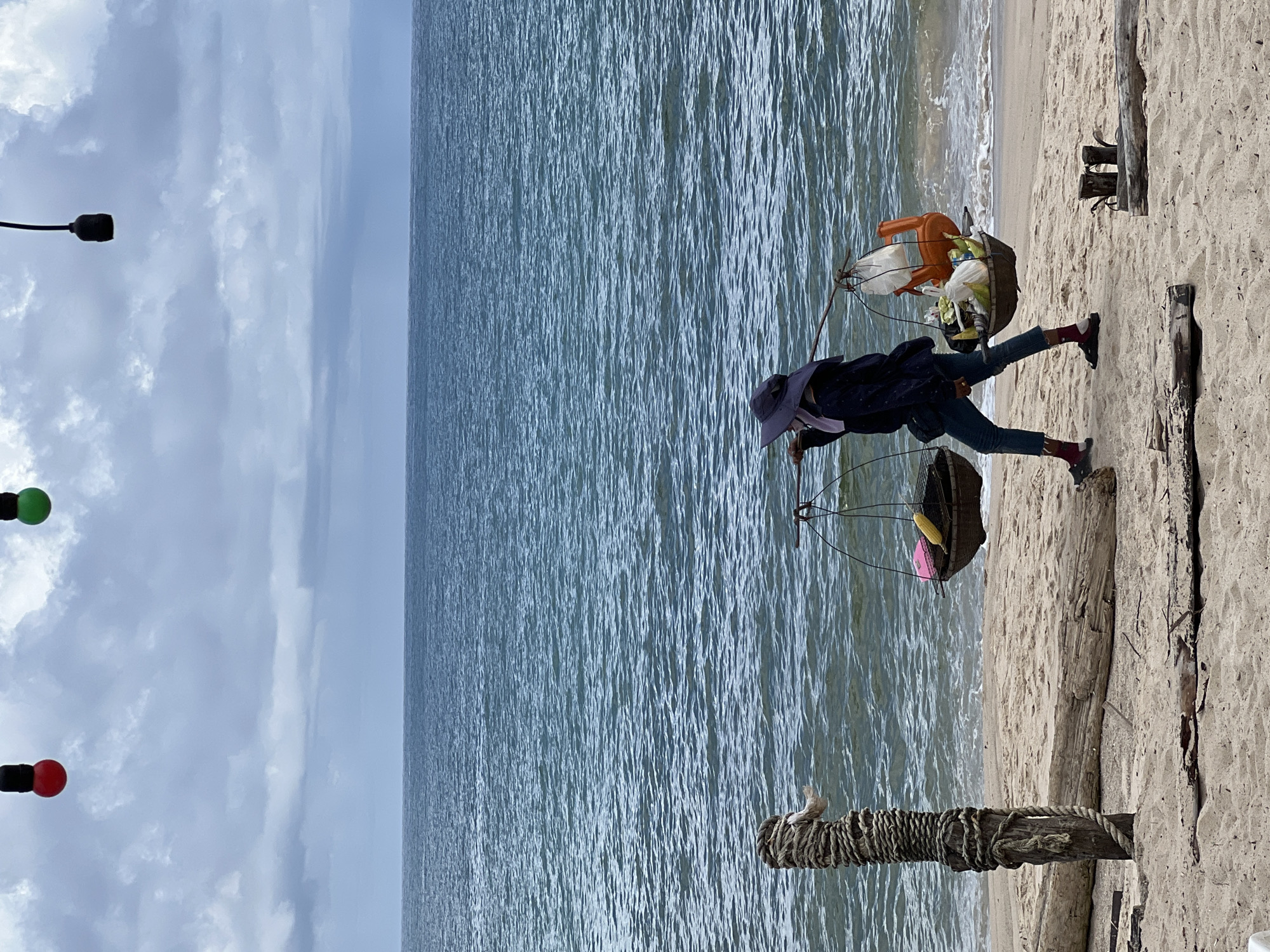 A person walks along a sandy beach carrying goods in baskets balanced on a pole over their shoulders. The person wears a wide-brimmed hat, a dark jacket, blue jeans, and dark sandals with red socks. In the left basket, a pink object and corn are visible. The right basket contains various items, including a small orange plastic chair and what appears to be more produce. The sea is visible behind the person, with choppy blue water under a cloudy sky. In the foreground, driftwood and a wooden post wrapped in rope lie on the sand. In the upper portion of the image, red and green lights are suspended against the sky. The scene is set in a natural environment, likely a beach.