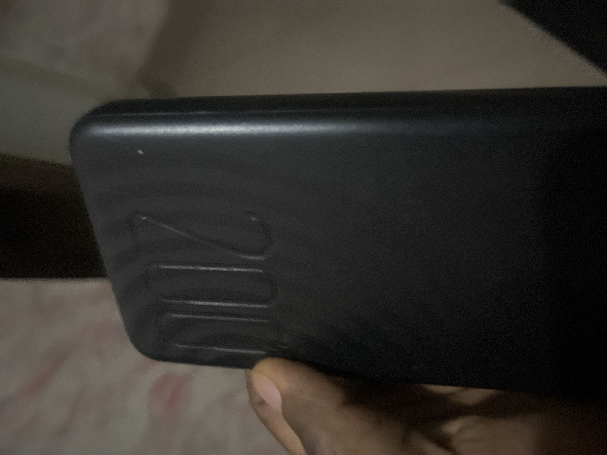 A person's hand is holding a black power bank with "200" embossed on it. The background is blurred and appears to be indoors with a textured surface, possibly a bedsheet, and a dark wall. The image is taken from a close-up perspective, focusing on the power bank and the hand. There is no identifiable information about the location in the city of Jalingo, Nigeria.