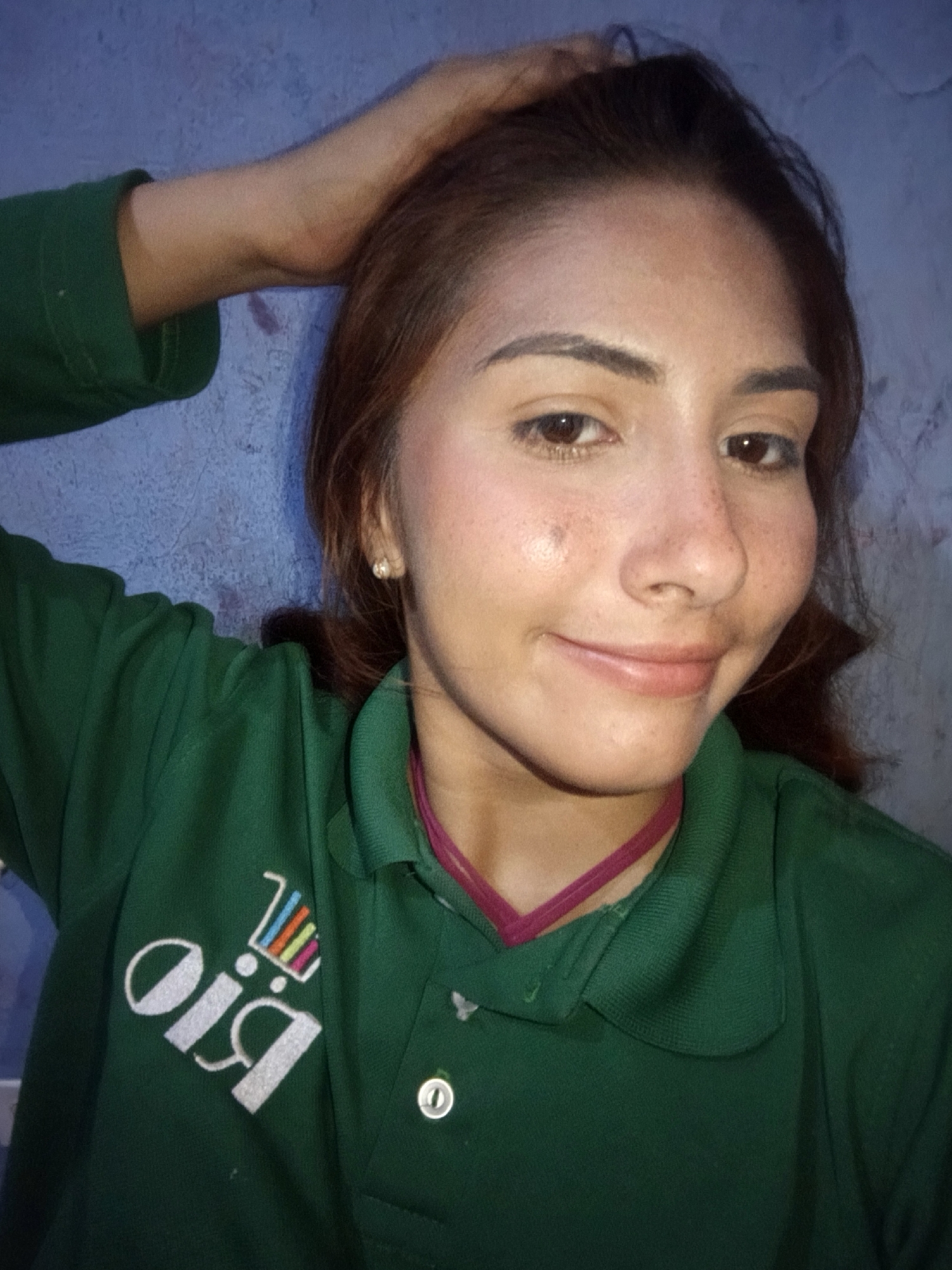 A young woman with light brown hair and a fair complexion is shown in a selfie. She is wearing a green collared shirt with a logo that reads "oiq" in white lettering, superimposed by a colorful shopping cart graphic. She has a pink ribbon choker around her neck. Her right hand is raised and resting on her head. She has a faint blush on her cheeks and small freckles across her nose and cheeks. The background is a blue, textured wall. The scene appears to be indoors. The location is not specified in the image.