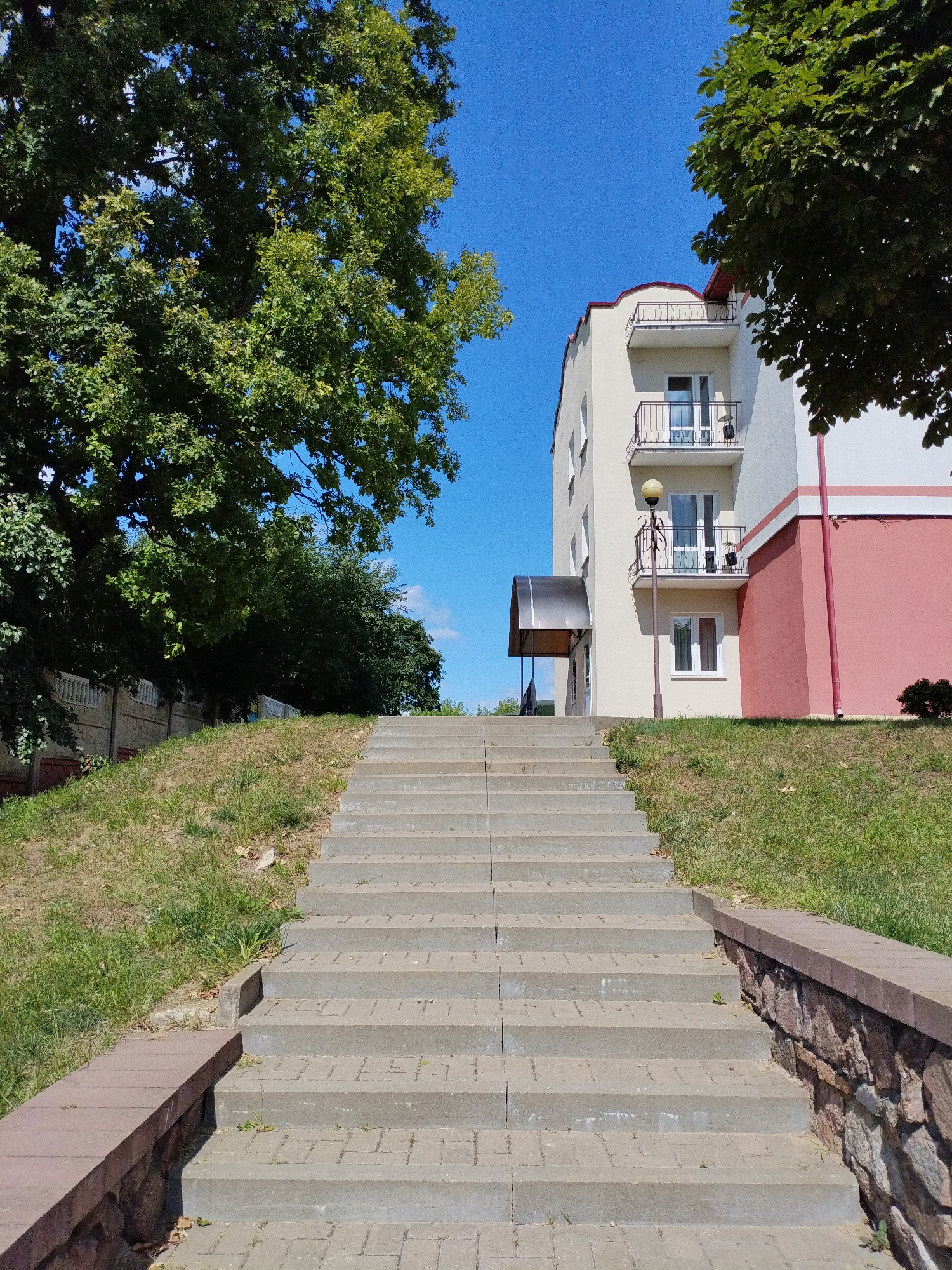 A concrete staircase leads up a grassy embankment towards a multi-story building. The building has cream-colored walls with pink accents and several balconies with black railings. A street lamp stands near the entrance. Large green trees frame the scene on the left and right. The sky is clear and blue. The location is Bialynichy, Belarus. There are no people visible in the image.