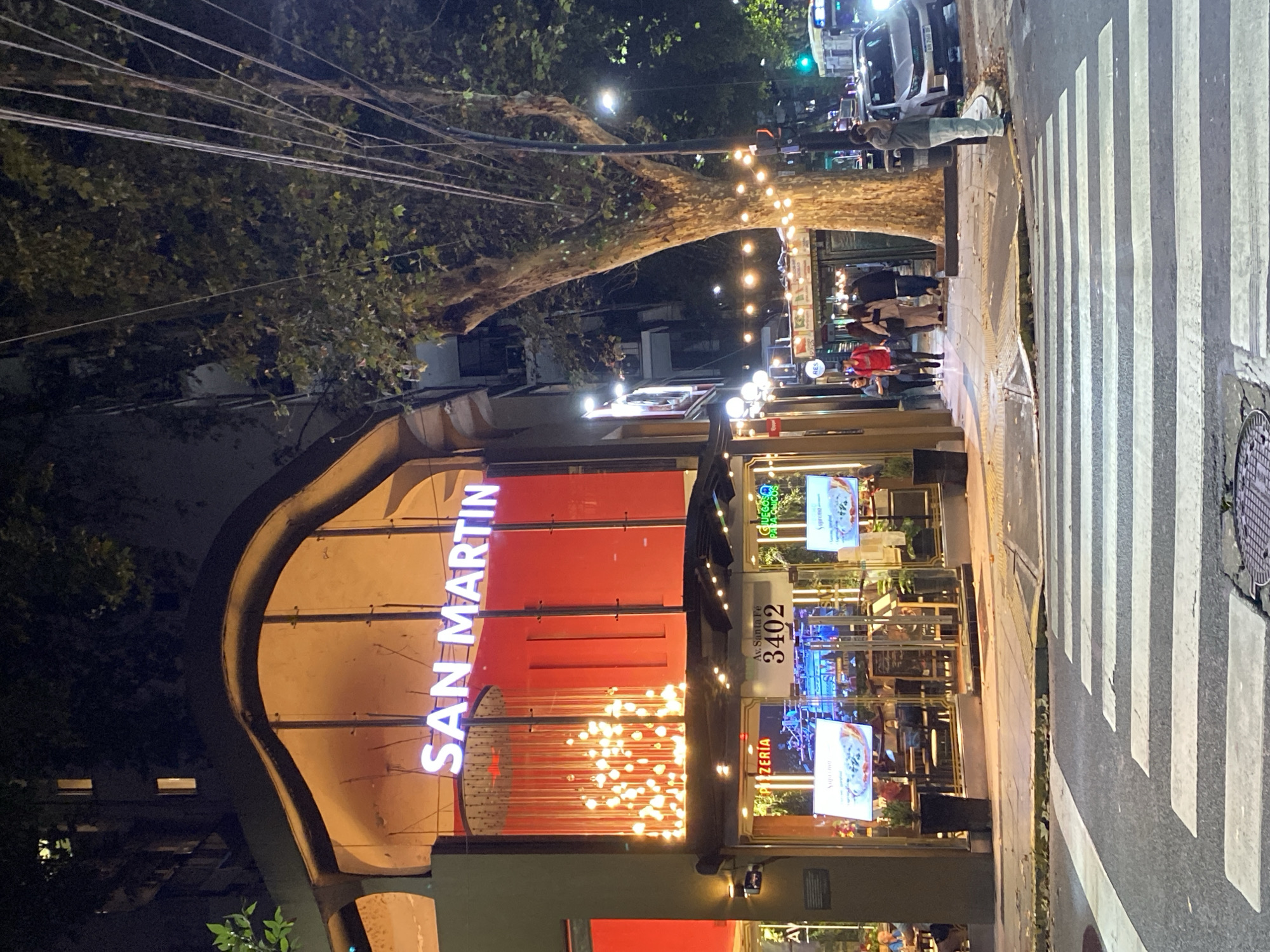 The image depicts an evening street scene in Buenos Aires, Argentina. The restaurant "SAN MARTIN" is prominently featured, with a glowing sign in capital letters, located at Av. Santa Fe 3402. The restaurant has an outdoor seating area with diners present. Above the seating area are strings of lights hanging from a large tree that extends over the street. People are seen walking along the sidewalk and waiting at a crosswalk. A vehicle is stopped at the crosswalk. The street is illuminated by streetlights and lights from the restaurant. The scene appears to be a typical evening in a bustling city neighborhood.