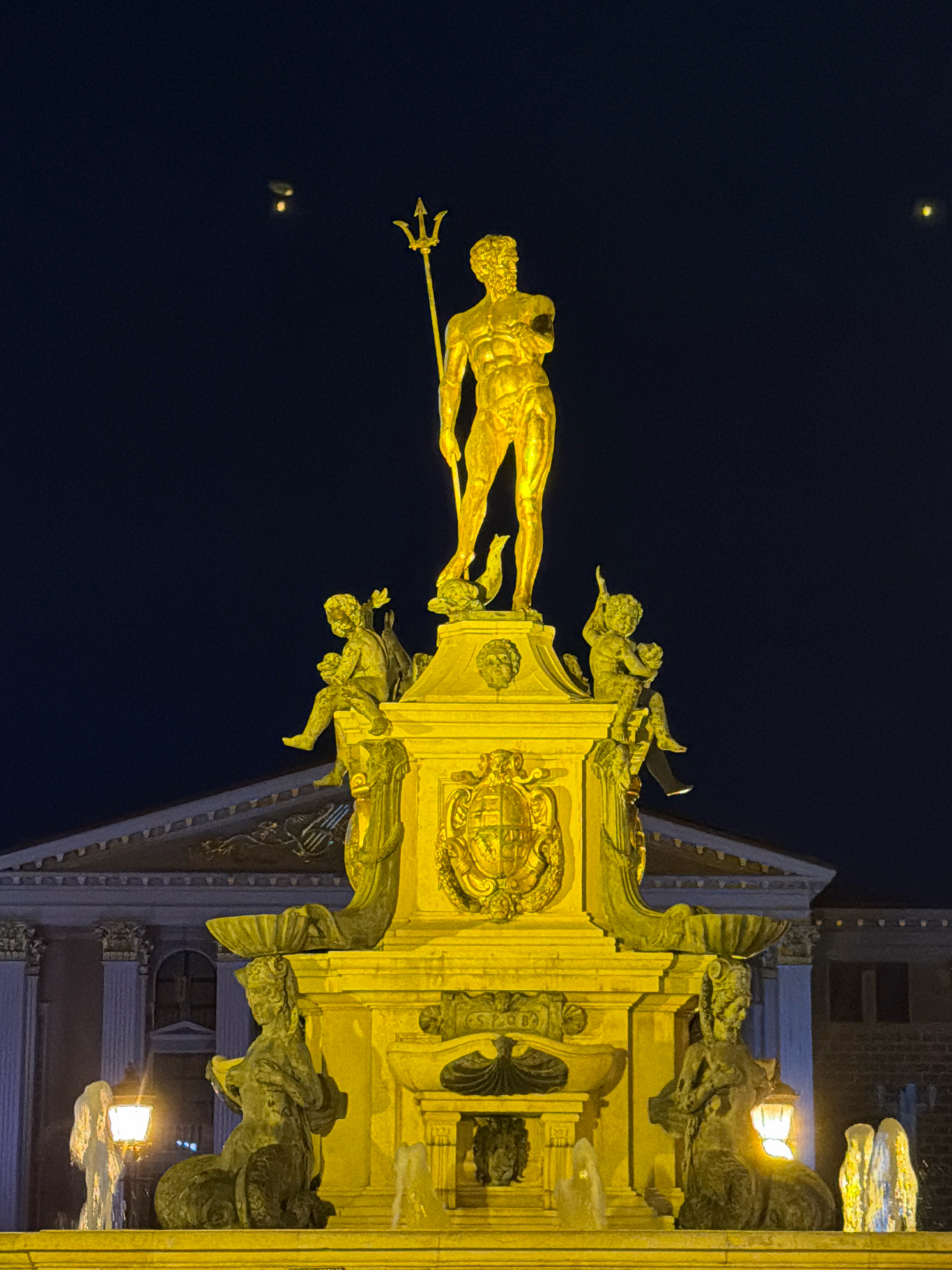 This is a nighttime photograph of the Neptune Fountain in Batumi, Georgia. The fountain is illuminated by yellow lights, highlighting its golden statue of Neptune at the apex, holding a trident. Below Neptune, on the pedestal, are four smaller figures, two on each side, depicting mythological beings, possibly nereids or tritons, with aquatic elements. The fountain itself is a multi-tiered structure with water features and ornate carvings. In the background, a building with classical architectural elements is visible, also illuminated. The sky is dark blue, with two distant lights, possibly aircraft or celestial bodies, visible in the upper left portion of the frame. The overall scene is dramatic and majestic, emphasizing the grandeur of the fountain.