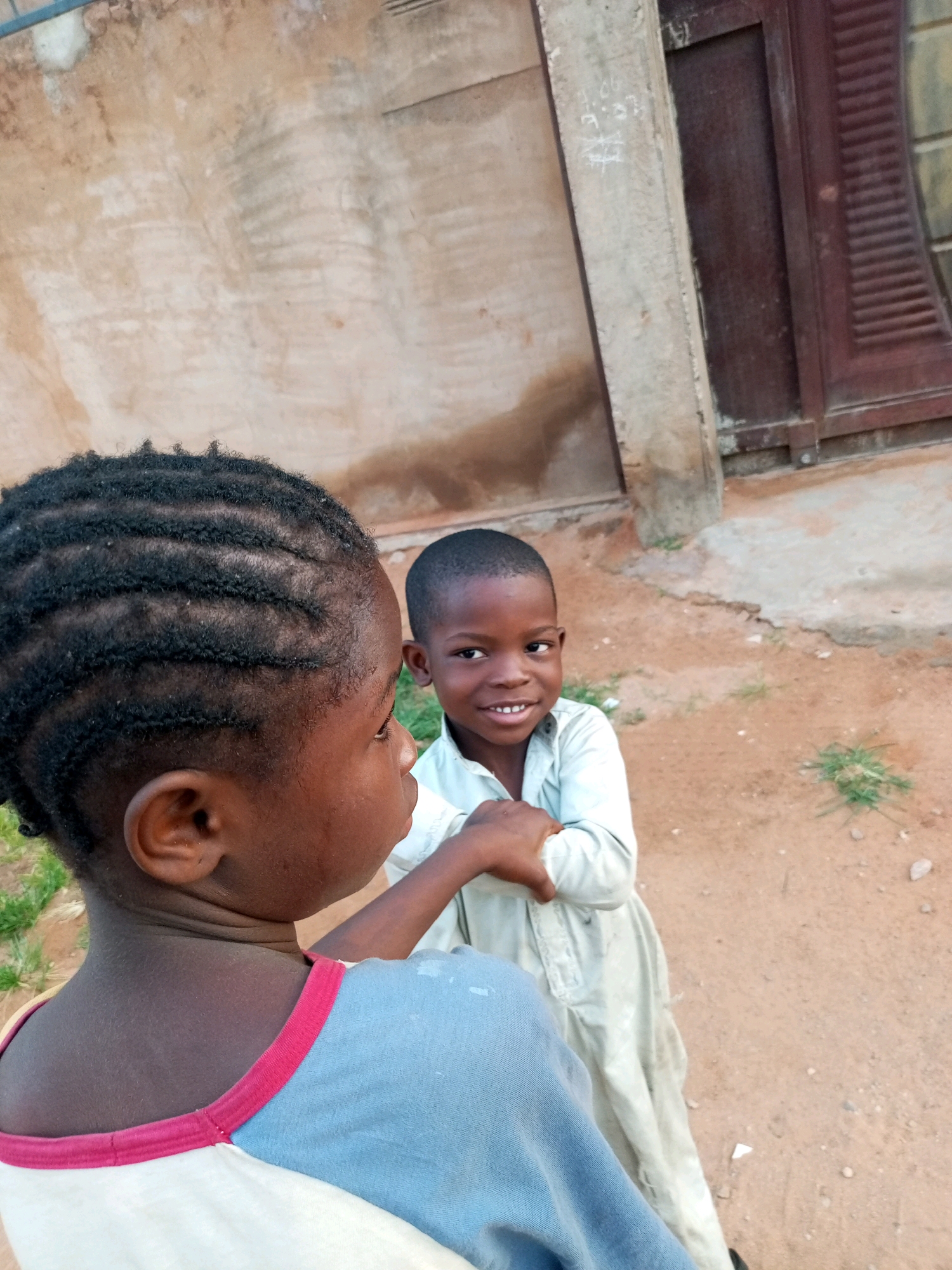 Two young children are interacting outdoors, possibly in an urban or semi-urban setting in Tempure, Nigeria. The child in the foreground, facing away from the camera, has their back and head visible. Their hair is styled in neat cornrows, and they are wearing a light blue shirt with a pink and white collar. The second child, facing the camera, is to the right of the first child. They have short hair and are wearing a light-colored, long-sleeved garment. Both children appear to be engaged in some form of interaction, with the second child's hand touching the first child's arm. The background consists of a textured, light-colored wall and a dark-colored shuttered door or window, suggesting a building structure. The ground is composed of reddish-brown soil with sparse patches of green grass. The lighting suggests it is daytime.