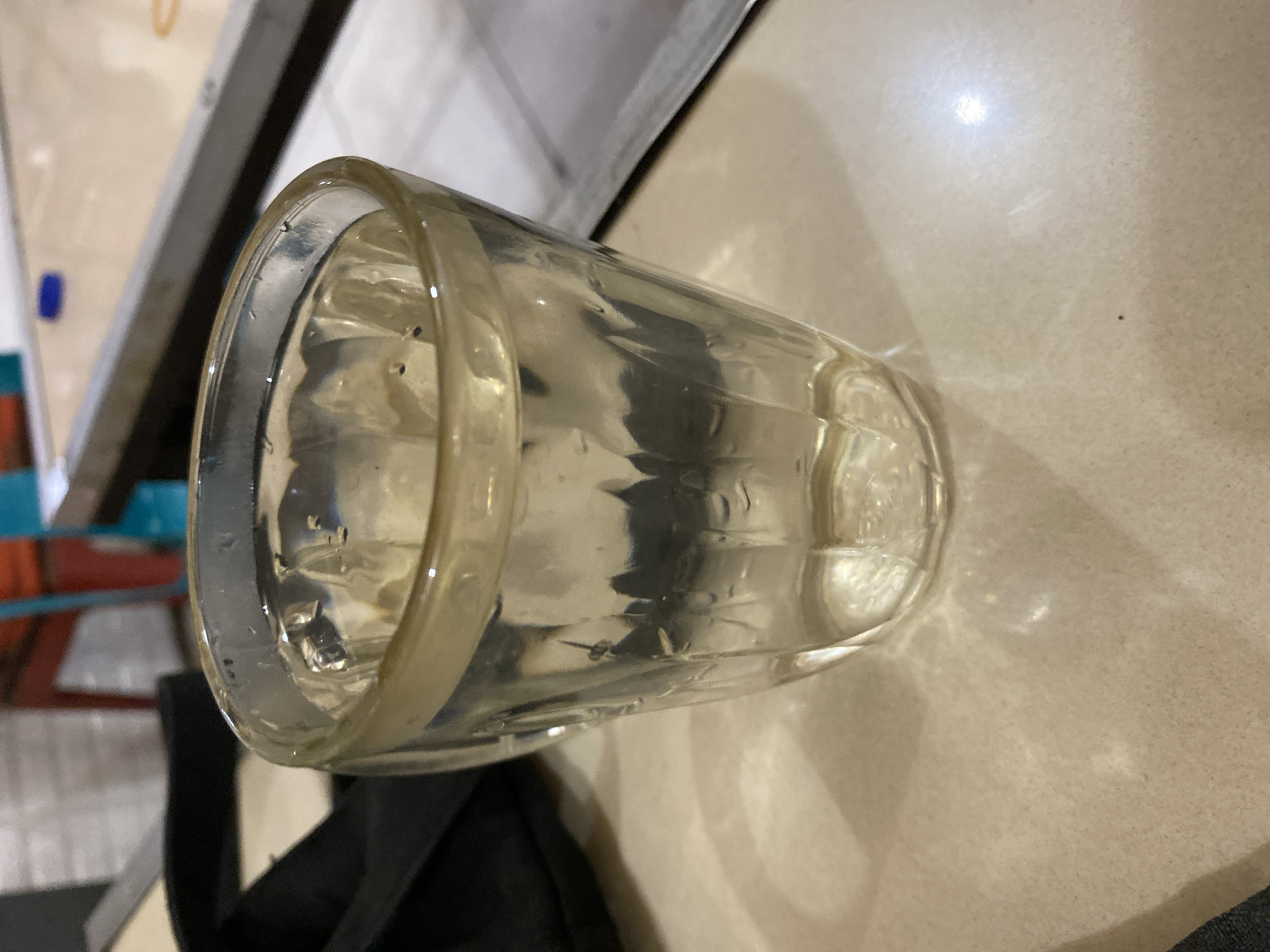 A glass of water sits on a light-colored tiled surface. The glass is clear with vertical ridges and has a slight yellow tint around the rim. It contains clear liquid with small bubbles and some darker particles floating on the surface. Behind the glass, a black bag is partially visible, and beyond that, blurred elements of a room with red and blue chairs and a long table with a light-colored surface are discernible. The lighting suggests an indoor environment, possibly a cafe or restaurant in Lhokseumawe, Indonesia.