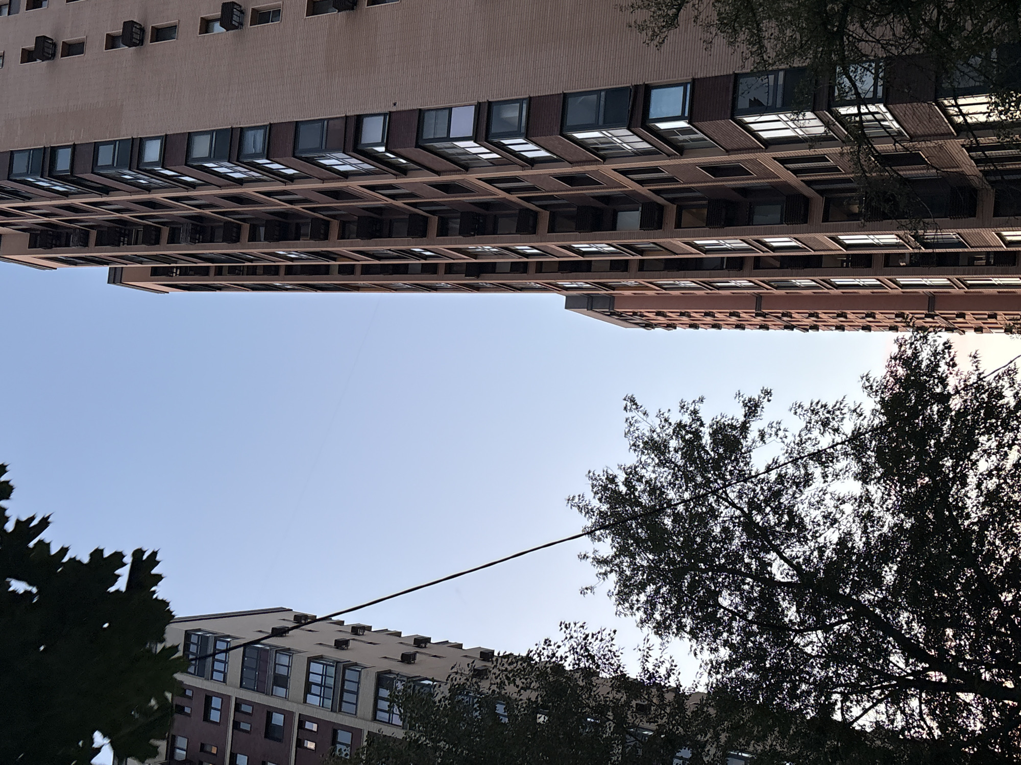 This media file captures a view looking up at two apartment buildings, with trees in the foreground and background. The buildings are tall and modern, with numerous windows arranged in a grid-like pattern. The facades are primarily a light brown or beige color, with darker accents around the window frames. Some windows appear to have individual air conditioning units mounted on the exterior.
The image is oriented such that the buildings are seen from a low angle, emphasizing their height. The sky is a clear, pale blue, suggesting a bright day. The presence of trees with dense foliage indicates that the scene is likely set during the warmer months, possibly spring or summer. The lighting suggests it is daytime, and given the gentle blue of the sky, it could be morning or late afternoon.
The composition creates a sense of urban environment, likely a residential area in Moscow, Russia. The view is a common urban scene, focusing on architecture and natural elements in close proximity. There is no discernible activity or event taking place; it is a still image of the architectural landscape. The overall mood is serene and observational, with no strong emotional cues conveyed. No text is visible in the image.