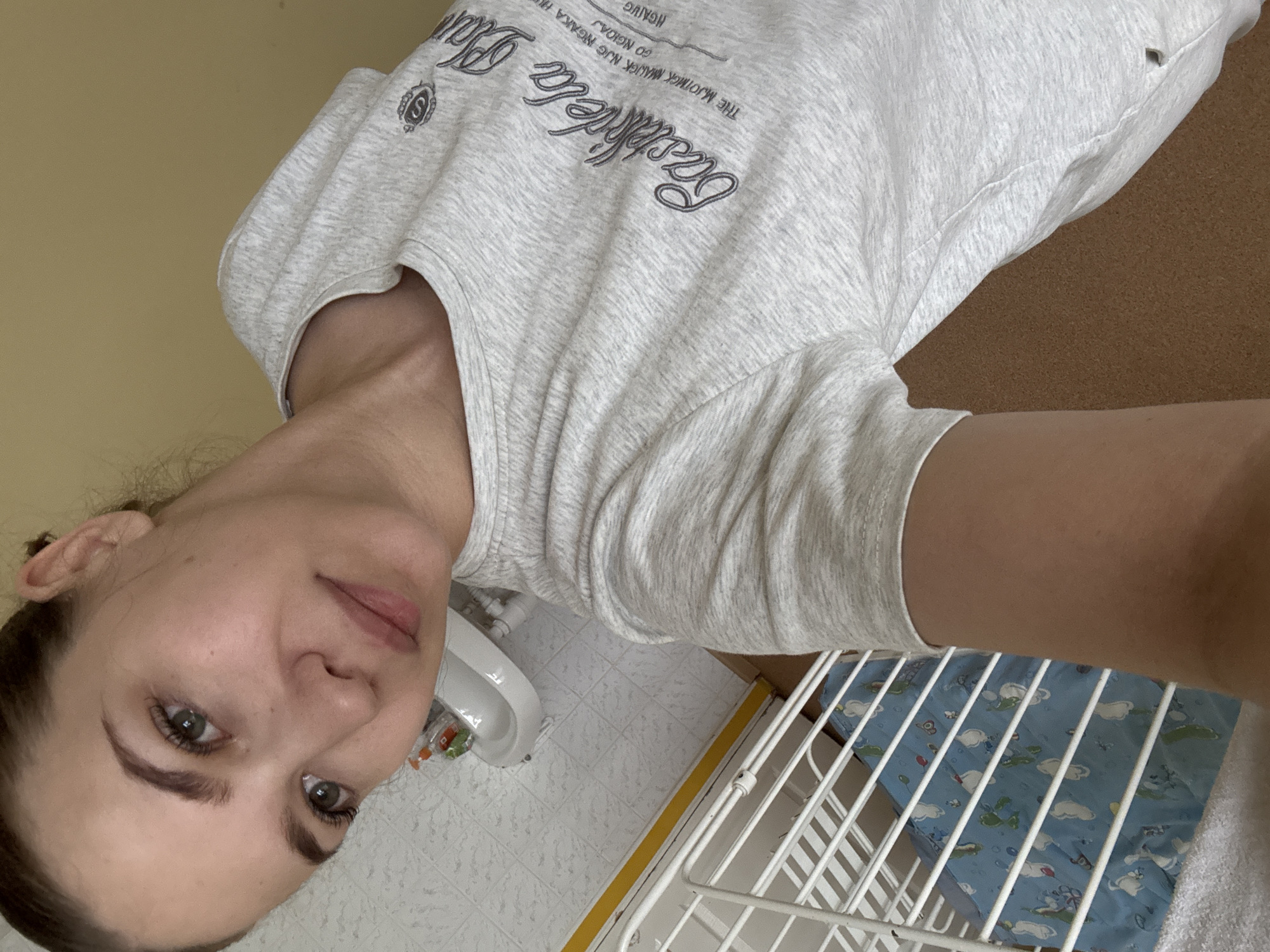 An adult female, appearing to be in her 20s to 30s, is captured in a selfie-style photograph indoors, in the city of Chaikovsky, Russia. Her dark brown hair is pulled back, and she has a fair complexion with minimal visible makeup, featuring defined eyebrows, green eyes, and light pink lips. A small mole or blemish is visible below her left eye. She wears a light grey marl short-sleeved t-shirt with dark grey cursive text on the chest, partially legible as "Castel dela", and additional smaller, partially visible, illegible text below it. A stylized 'S' emblem is present above the main text and on her left shoulder.
The background depicts a domestic interior. To the left, a section of white square-tiled flooring with dark grey grout lines is visible. Adjacent to this tiled area, a white metal structure, resembling a baby crib or playpen, contains a blue mat or sheet adorned with cartoon animal figures, predominantly white and green. A white towel is draped over the lower part of the metal structure. Above the tiled floor, a small white wall-mounted basin sink is present, with visible plumbing pipes underneath. An orange and clear plastic bottle, along with a smaller green object, rests on the sink's edge. A thin yellow horizontal stripe delineates a boundary between the tiled floor area and an adjacent section of light brown, textured flooring, possibly cork. The wall in the background is a plain, solid light yellow-beige color.