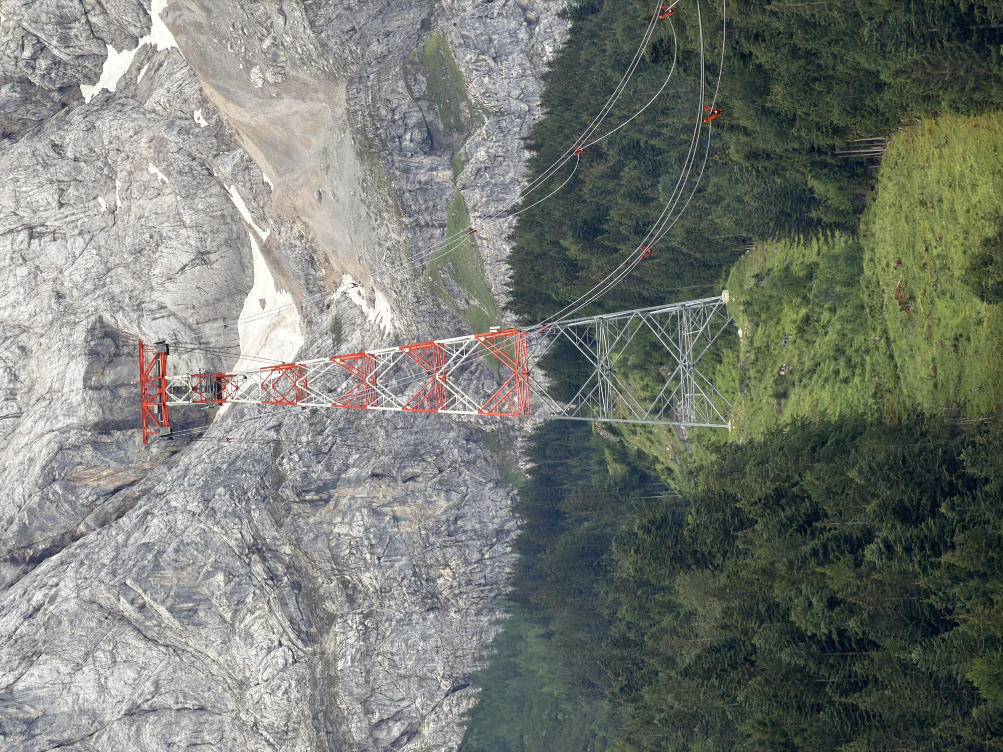 This is a medium shot, eye-level, vertical photograph of a red and white pylon for a cable car, set against a backdrop of a rugged, grey mountain face. The pylon is positioned in the center of the frame, extending from the lower portion upwards. It is a lattice structure with red crossbeams and white supports. Cables extend from the top of the pylon towards the right side of the frame, disappearing into the distance.
Below the mountain face, a dense forest of dark green coniferous trees covers a steep, grassy slope. The trees are thick and lush, suggesting a high altitude or alpine environment. The slope is uneven, with patches of green grass and some rocky outcrops.
In the upper portion of the image, the mountain face dominates the background. It is a sheer, rocky surface with visible strata and texture, suggesting a geologically ancient formation. There are patches of snow or ice visible on the upper slopes of the mountain, indicating a cold climate or high elevation.
The overall scene depicts a dramatic landscape where man-made infrastructure, the cable car pylon, is integrated into a natural, mountainous environment. The location is identified as Grainau, Germany, which is known for its proximity to the Bavarian Alps and its popular tourist attractions, including cable cars that ascend the mountains.