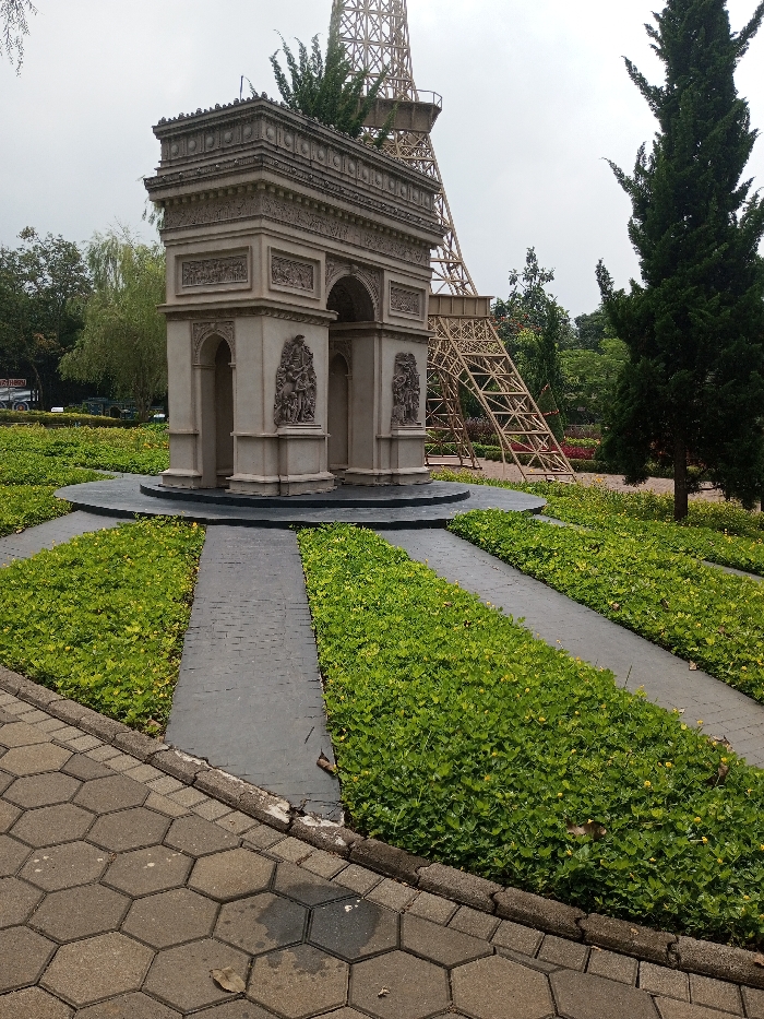 The image depicts an outdoor park setting in Ciherang, Indonesia, featuring miniature architectural replicas under an overcast sky. In the central foreground, a detailed, light-colored replica of the Arc de Triomphe stands on a dark circular base. This structure exhibits classical architectural details, including carved reliefs and multiple arched passages. To the right and behind the Arc de Triomphe, a large, skeletal, light brown lattice structure, representing a section of the Eiffel Tower, is prominently visible. Its lower framework extends into the midground.
The ground around the Arc de Triomphe is landscaped with dark gray paved paths radiating outwards, alternating with dense sections of vibrant green, low-lying ground cover. In the immediate foreground, a pathway constructed from light gray hexagonal pavers runs horizontally across the bottom of the frame, separated from the landscaped area by a concrete curb. Scattered across the scene are various trees: a tall, slender evergreen with dark green foliage is on the far right, and a lighter green weeping tree is visible on the left. The sky is a uniform, light gray, indicating an overcast condition. No individuals or overt actions are present in the scene.