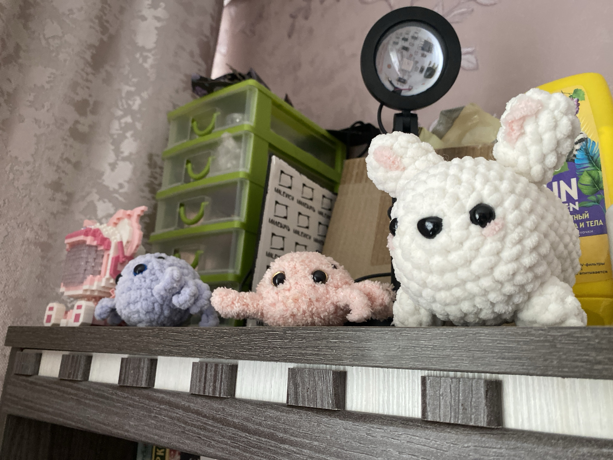A close-up shot captures a collection of decorative items arranged on a dark wooden shelf. The primary subjects are three crocheted plush toys: a fluffy white bunny with pink inner ears sits prominently on the right, facing slightly left. In the center is a smaller, pink octopus-like plushie, and on the left, a light purple, blob-shaped creature with blue eyes. All three plushies feature large, dark, bead-like eyes.
Behind the plush toys, on the left side of the image, is a green plastic organizer with three drawers. A small black ring light, reflecting its LED bulbs, stands behind the white bunny. Partially visible on the far right, behind the bunny, is a bright yellow plastic bottle. To the left of the purple plushie, a pink and white building toy, constructed from small blocks, is displayed. The wooden shelf itself has a decorative edge, with alternating vertical strips of dark wood and white. The background consists of a light-colored wall with a textured, possibly abstract or floral, pattern. The scene is brightly illuminated, suggesting an indoor setting.
Visible text on the yellow bottle includes Russian words such as "ный", "и тела" (and body), "У-фильтры" (U-filters), and "вляются". A piece of paper leaning against the green organizer also shows fragmented black text and squares.
